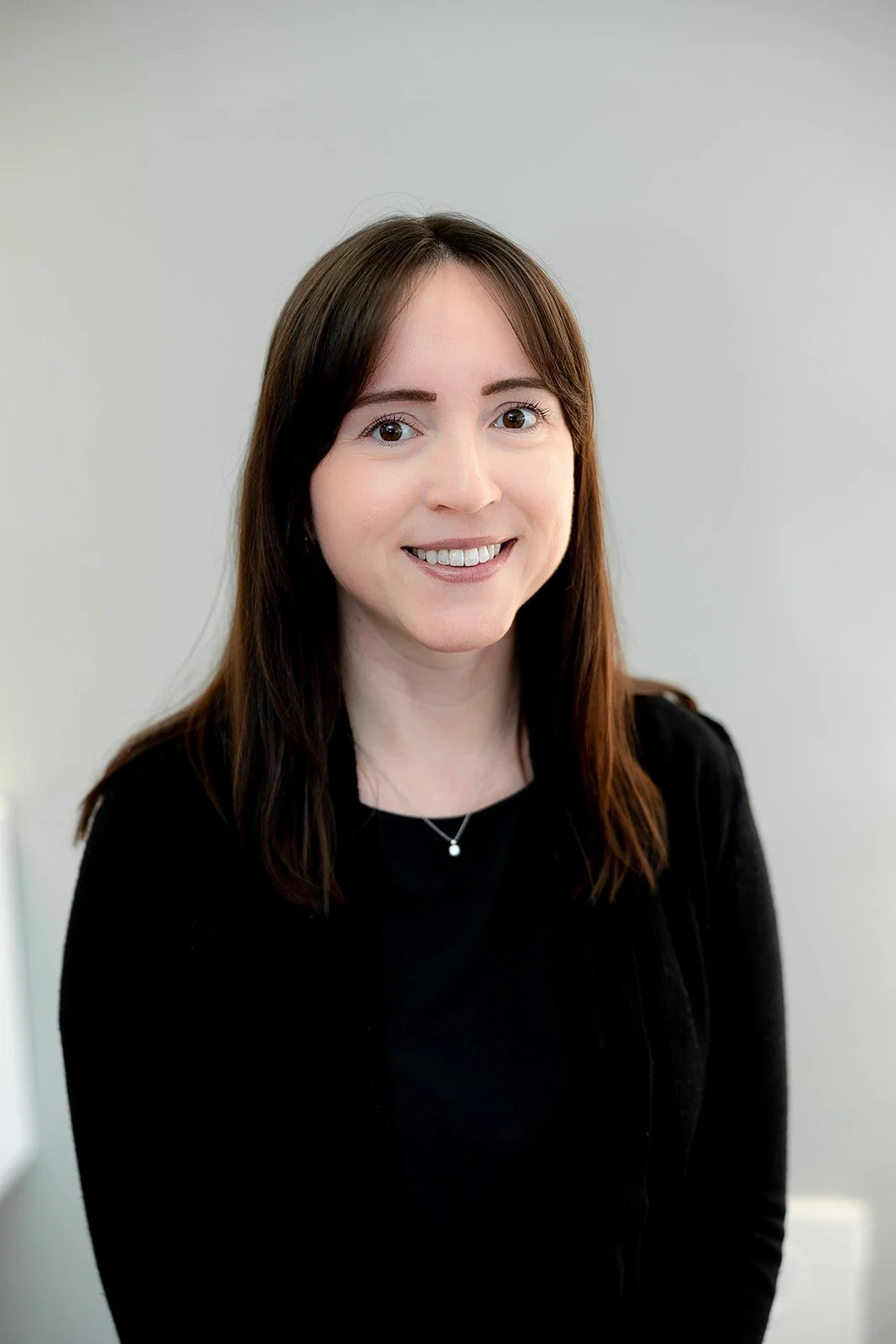 Portrait of a woman with shoulder-length dark brown hair, wearing glasses, a black top, a light-colored cardigan, and a gold necklace, standing against a plain light gray background and smiling. staff at peterborough optometric