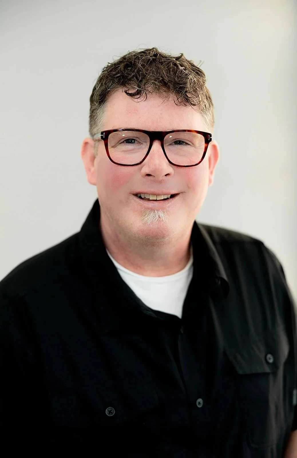 A man standing in front of a display of eyeglasses in an optical store, wearing a green polo shirt with the logo 'Peterborough Optometric'.