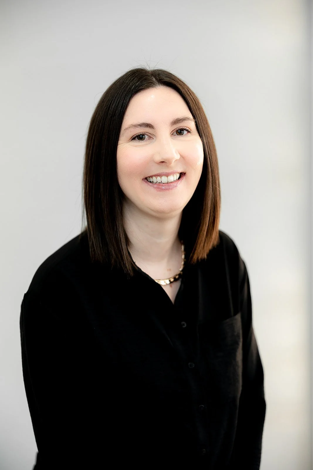 A woman with shoulder-length brown hair, blue eyes, wearing a black top and a burgundy blazer, smiling against a plain white wall background.