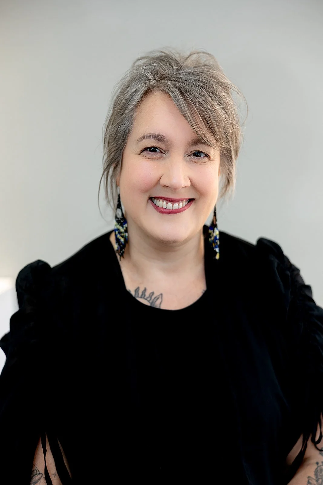 A smiling woman with gray hair and glasses, wearing a patterned black blouse and a white cardigan, standing against a plain light-colored wall. staff at peterborough optometric