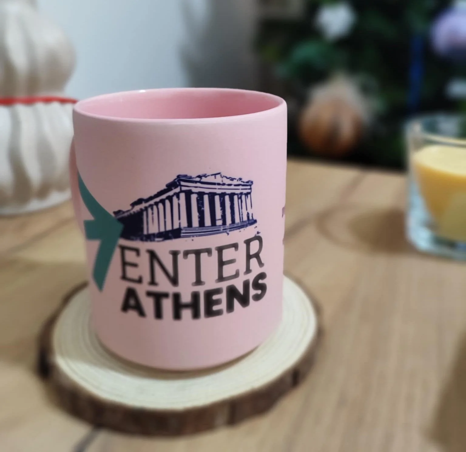 Pink mug featuring the Parthenon and text 'ENTER ATHENS' on a wooden coaster on a wooden table, with a blurred candle and other objects in the background.