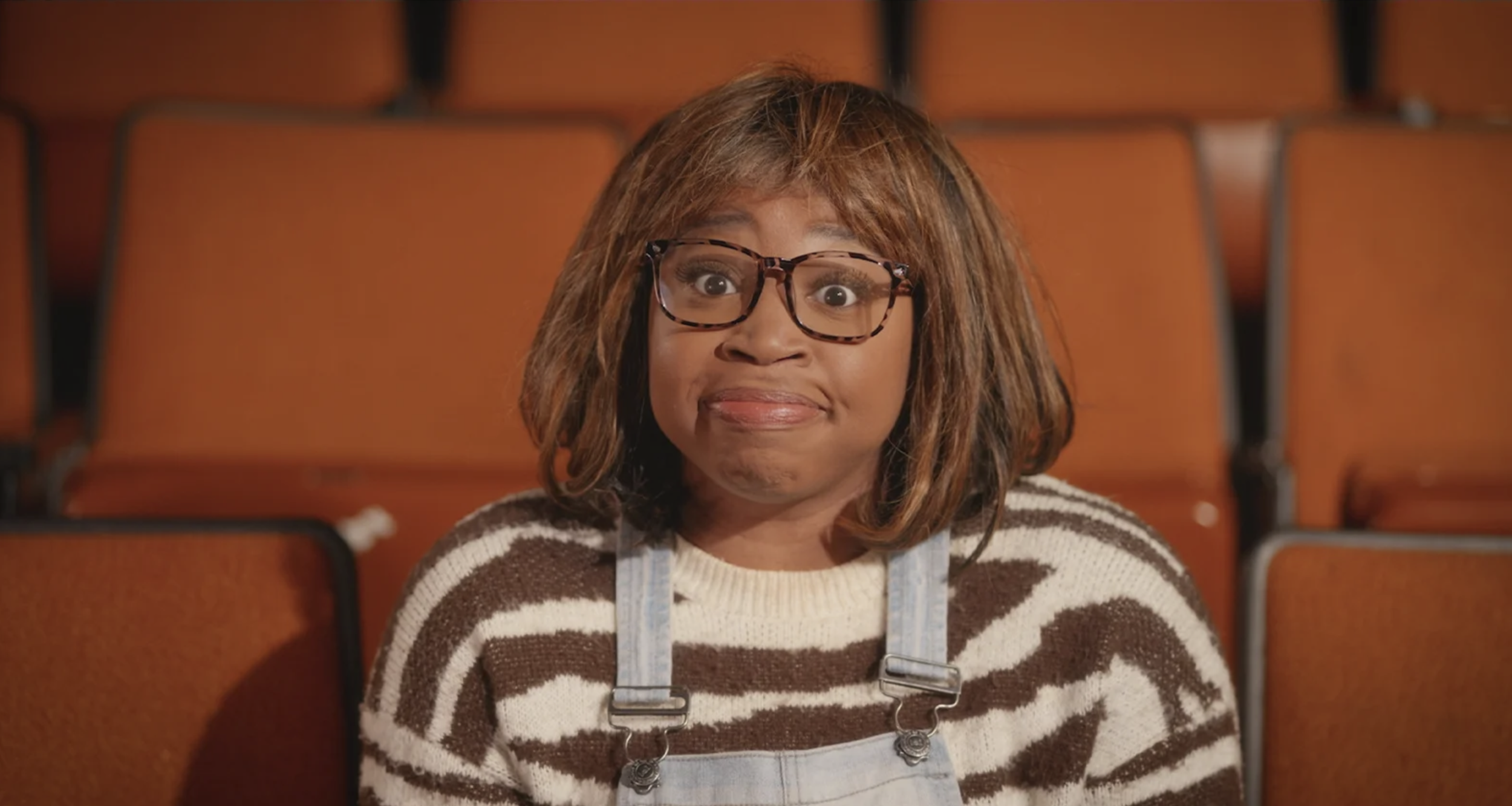 Woman with glasses and shoulder-length hair sitting in an empty theater, wearing a striped sweater and overalls, making a confused or unimpressed facial expression.