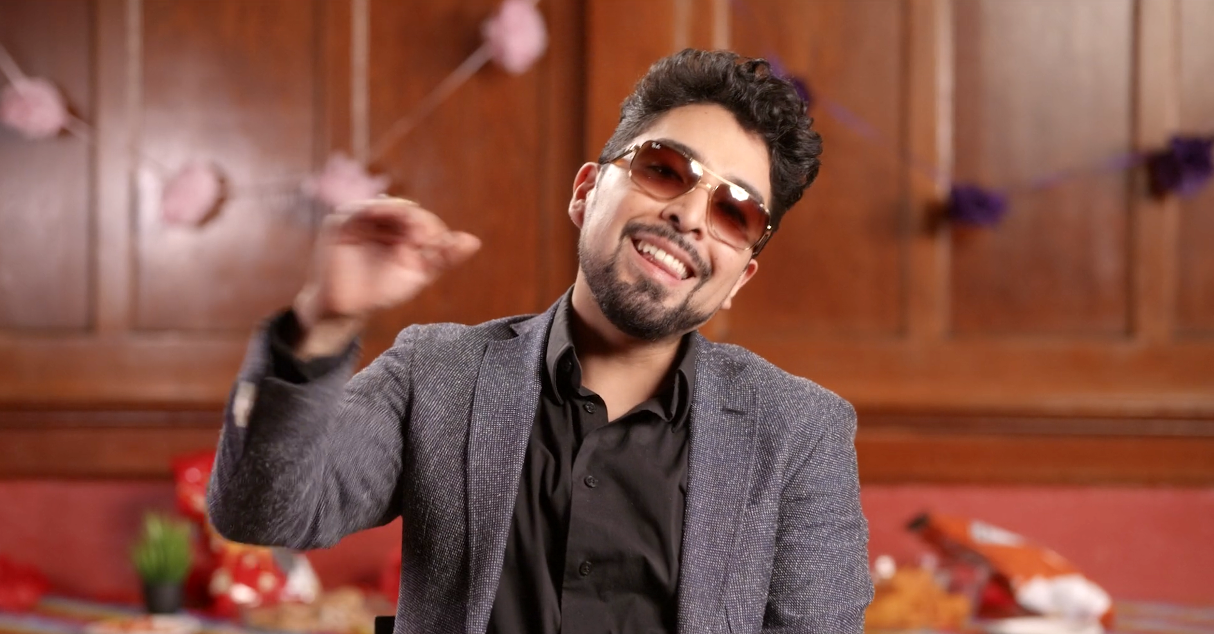 A man wearing sunglasses, a black shirt, and a gray blazer smiling and gesturing with his right hand.