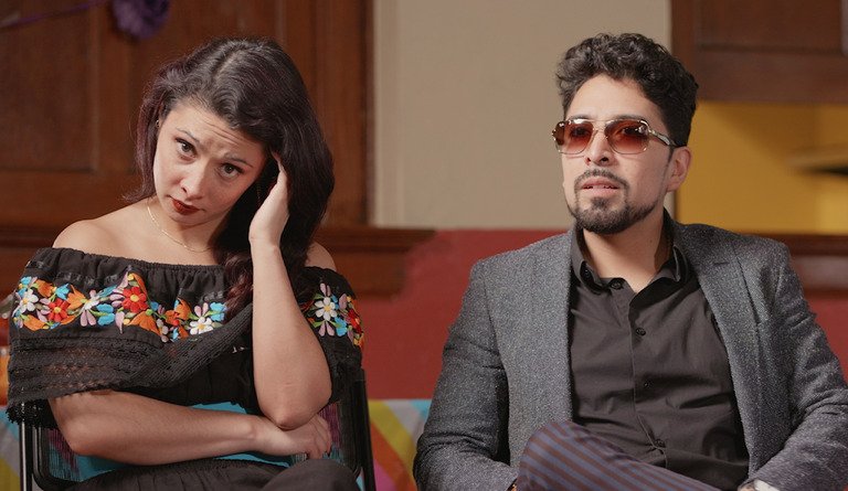 A woman and a man sitting indoors. The woman appears confused or concerned, with her hand on her head. She is wearing an off-the-shoulder black top with colorful floral embroidery. The man has a serious expression, wearing sunglasses, a dark button-up shirt, and a gray blazer.