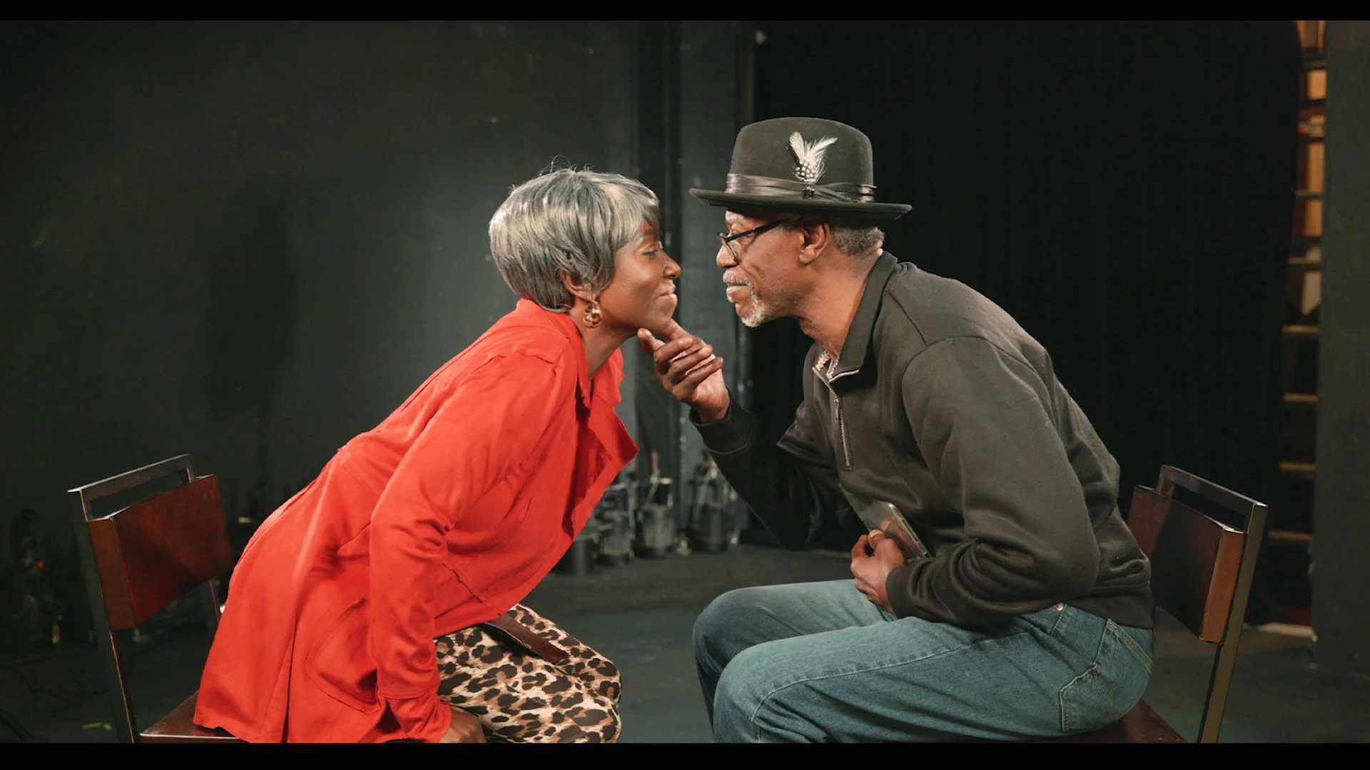 An elderly woman and man sitting closely on chairs, touching noses and gazing at each other affectionately in a theater or studio setting.
