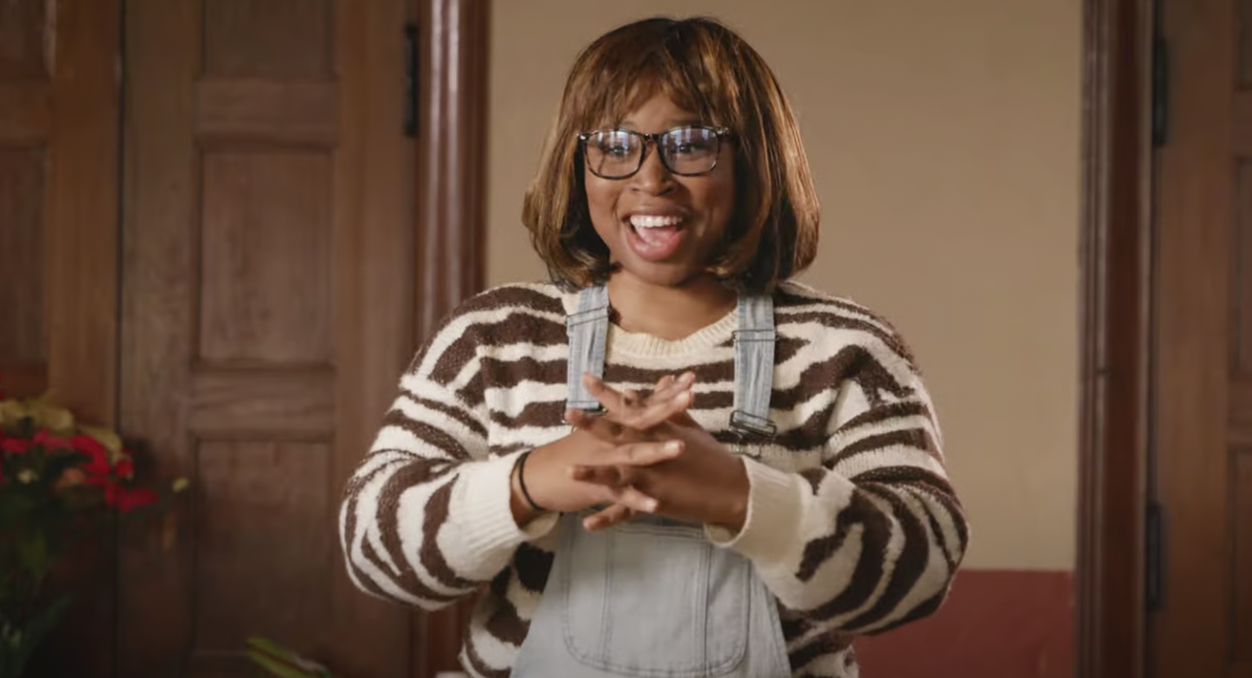 A woman with short brown hair and glasses, smiling with her mouth open, wearing a striped sweater and denim overalls in a warmly lit room.