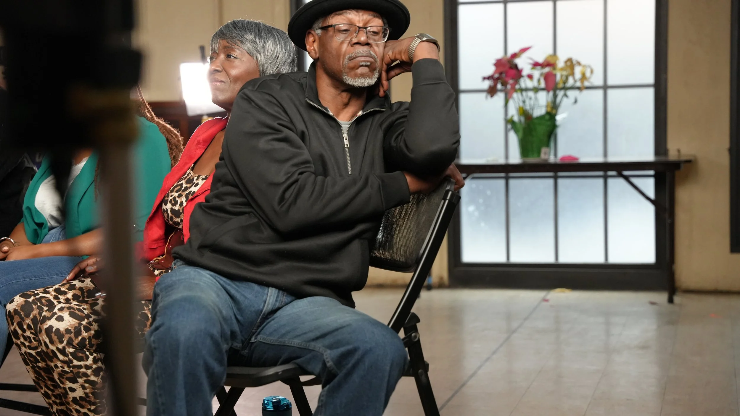 An elderly man with glasses and a hat sitting with his eyes closed and resting his head on his sitting arm in a room with a large window and a flower pot on the windowsill.