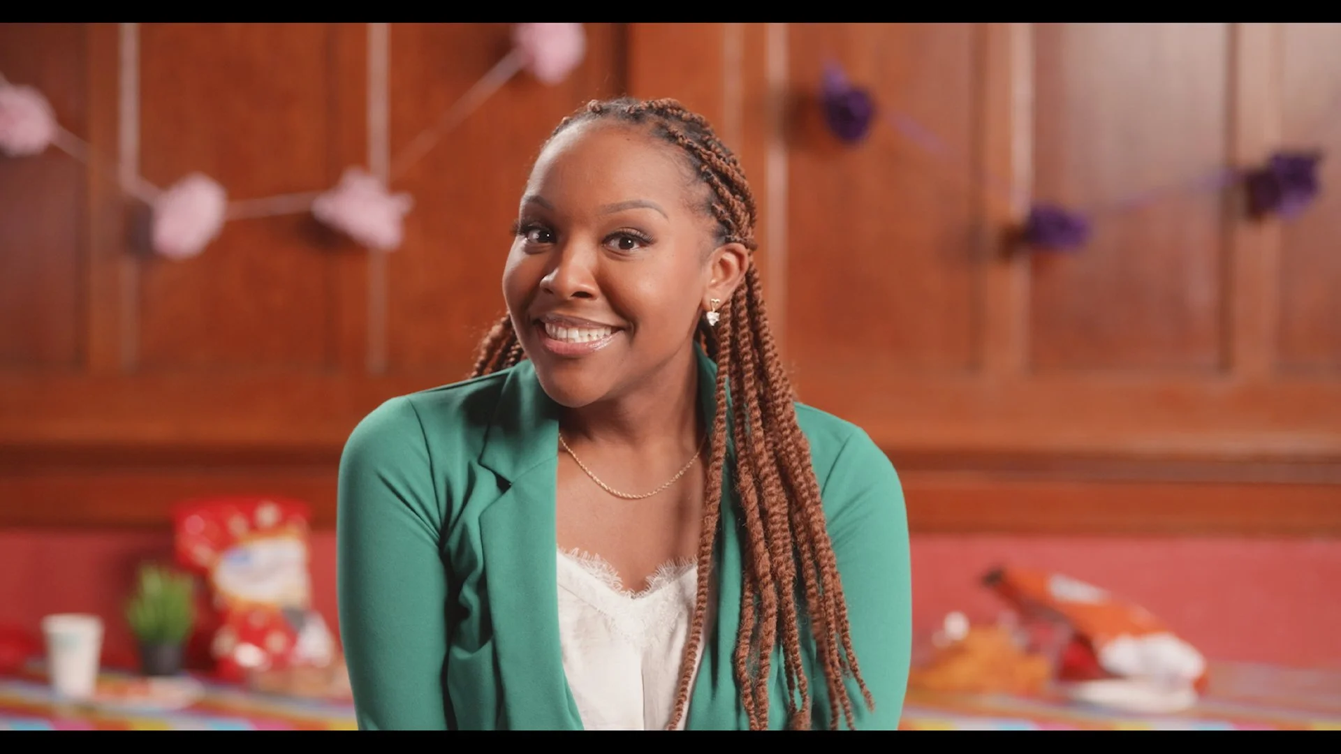A woman smiling sits in a colorful restaurant or cafe setting with a wooden wall decorated with purple paper flowers in the background.