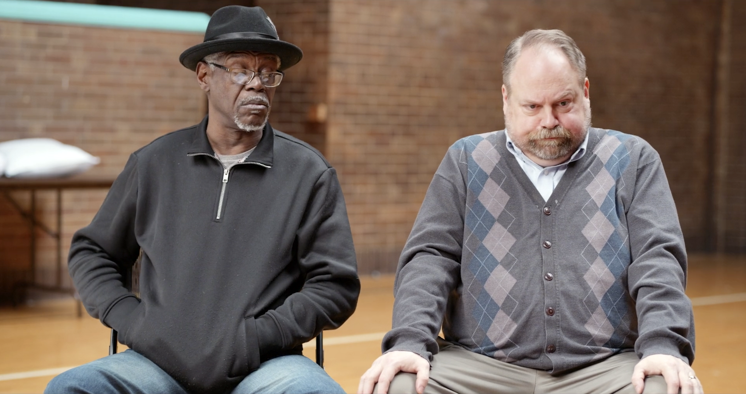 Two men sitting in a room with brick walls, one black man wearing glasses, a black hat, and a black zip-up jacket, the other white man with a beard wearing a gray argyle cardigan and a light blue shirt, both appearing serious or contemplative.