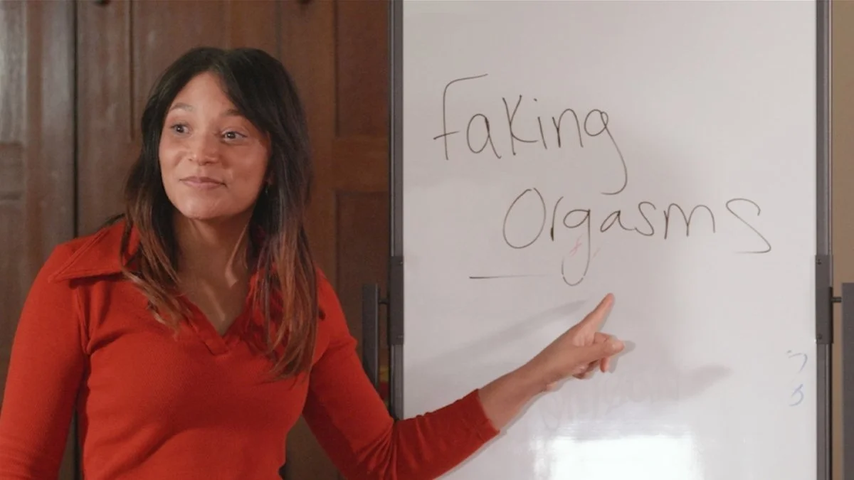 A woman in a red shirt points to a whiteboard with the words "faking orgasms" written on it.