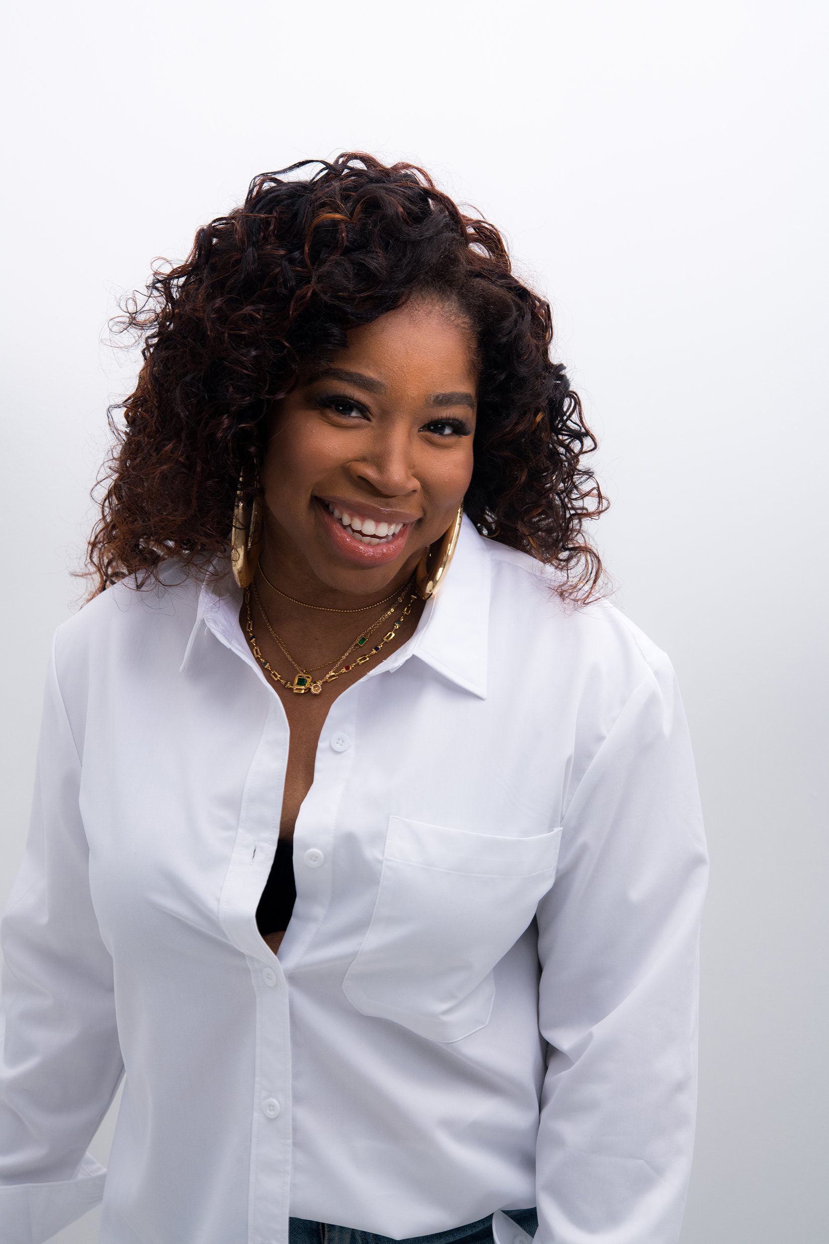 A woman with curly hair, wearing a white button-up shirt, gold hoop earrings, and layered gold necklaces, smiling at the camera against a plain white background.