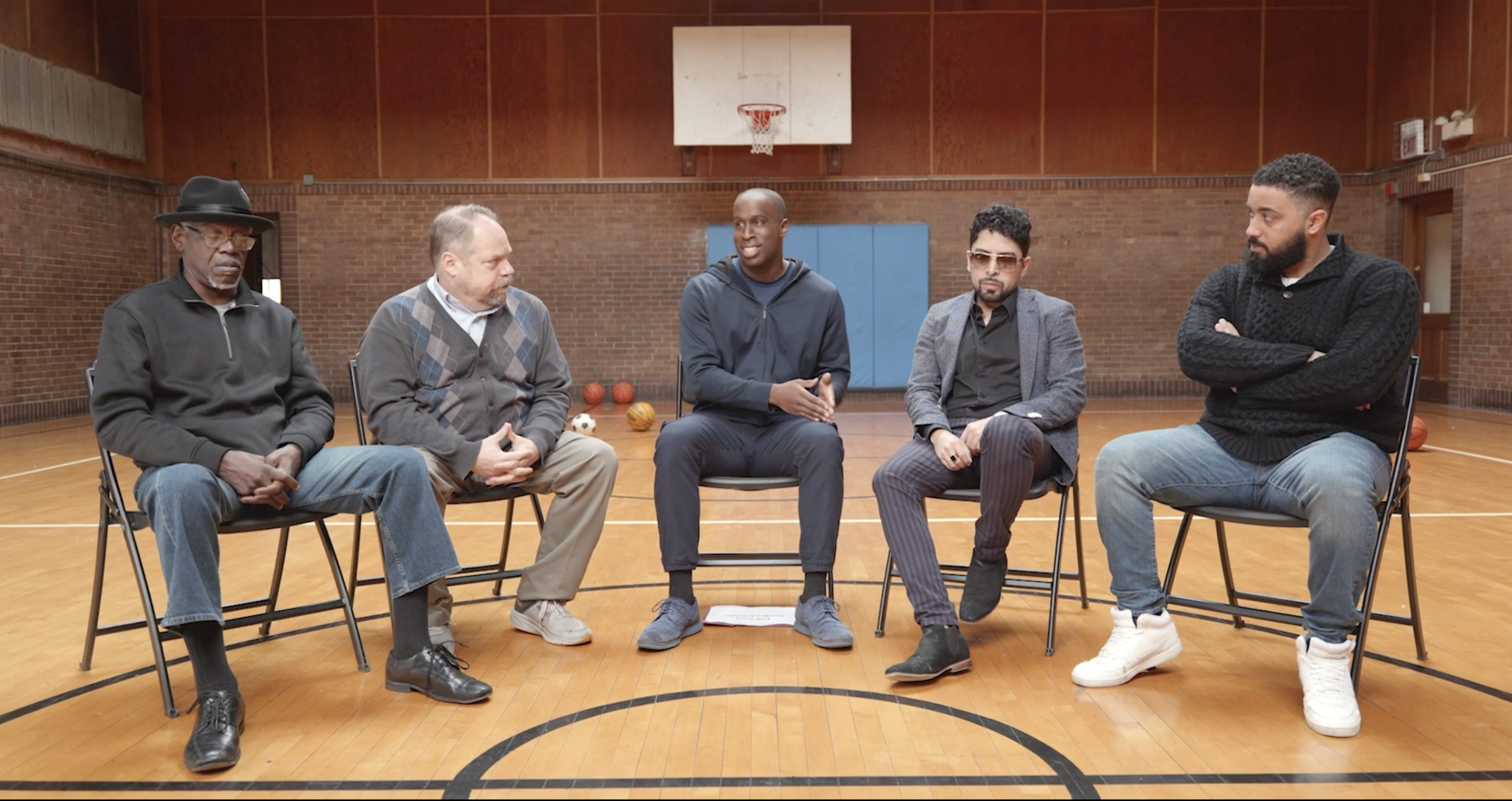 Five men sitting on chairs in a gymnasium during a communication workshop, with basketballs on the floor behind them.