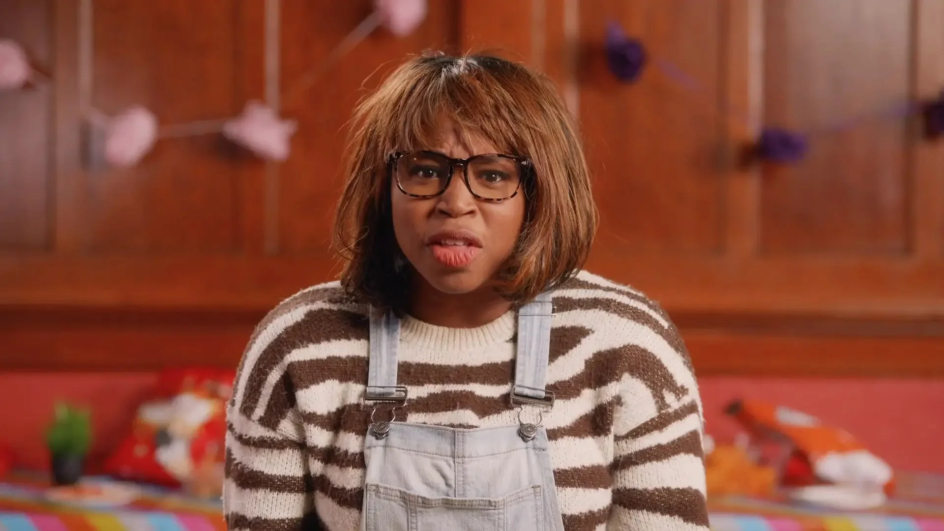 A woman with glasses and shoulder-length brown hair wearing a brown and white striped sweater and light-colored overalls, looks confused or annoyed in a room with wooden paneling and scattered items in the background.