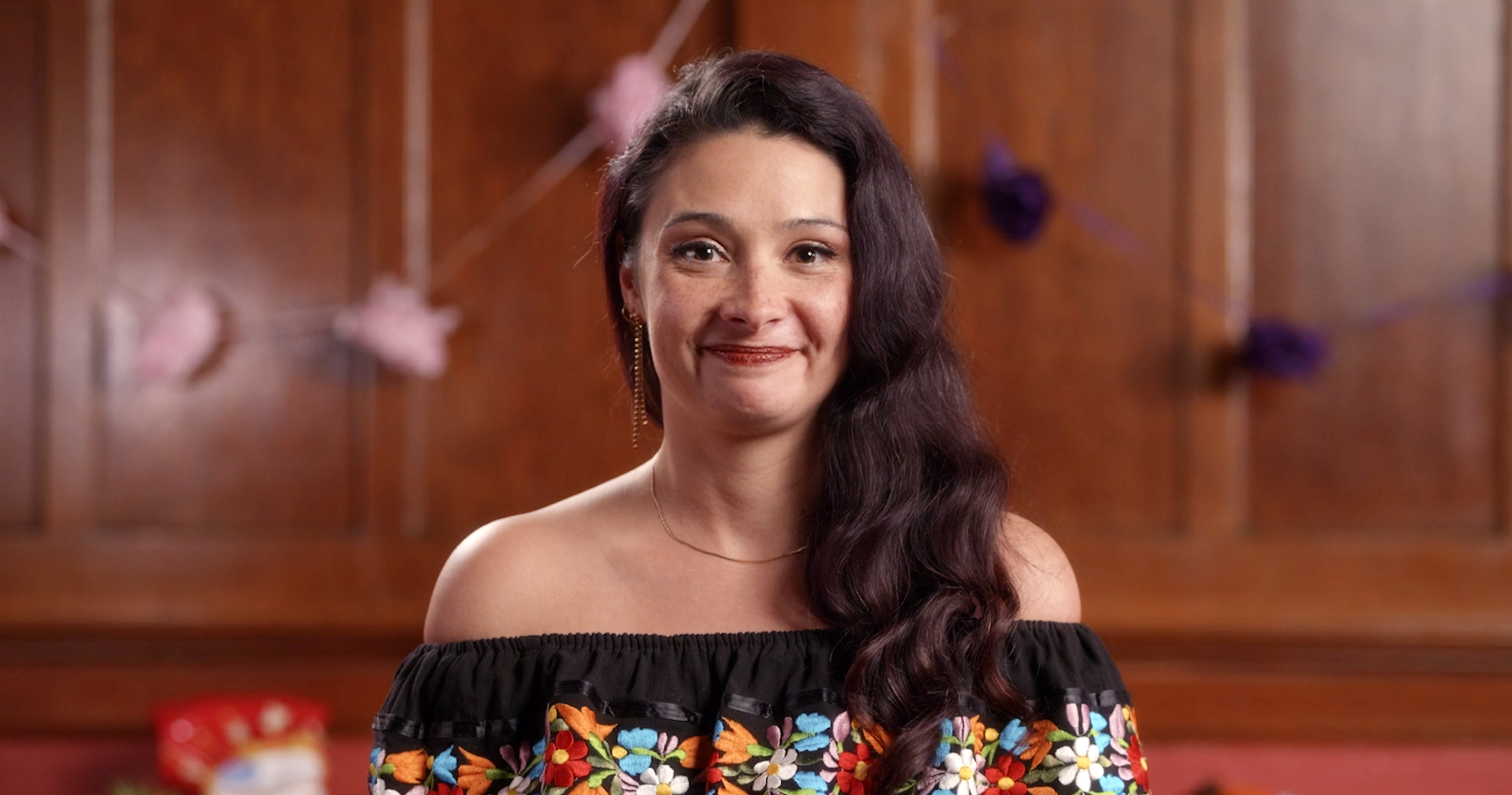 A woman with long, dark wavy hair wearing an off-the-shoulder black dress with colorful floral embroidery, smiling in front of a wooden background decorated with pink and purple flowers.