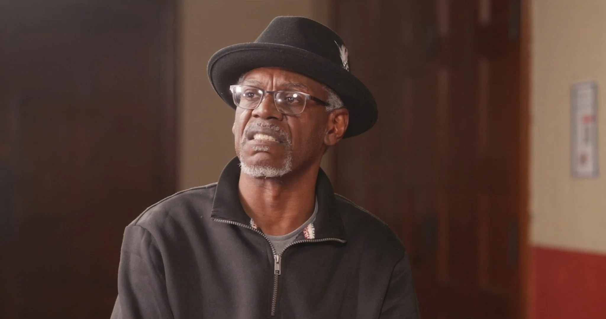 An older man with glasses and gray hair, wearing a black fedora hat, a black zip-up jacket, and a gray shirt, looking confused or concerned indoors with a wooden door and beige walls in the background.