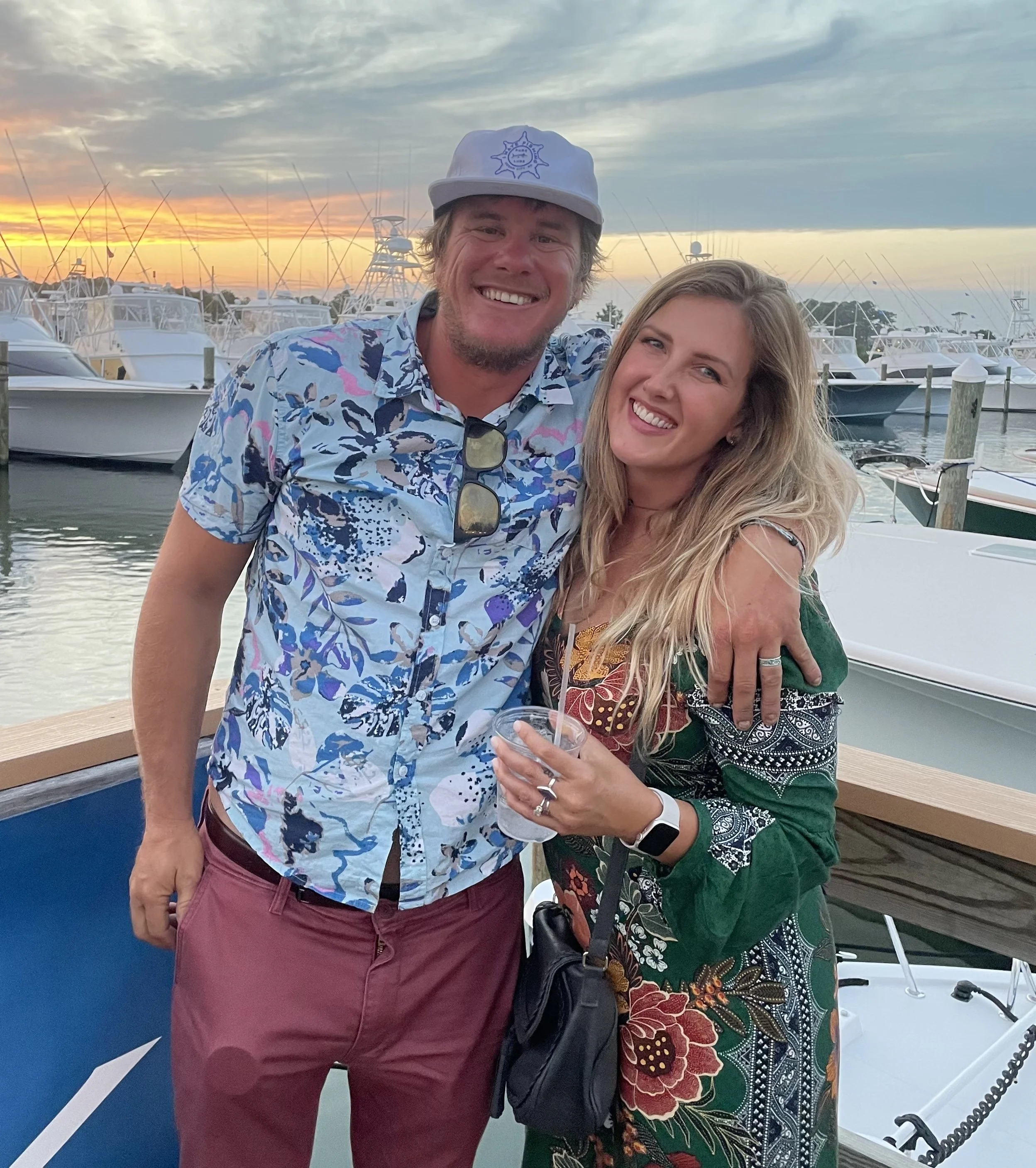 A smiling man and woman embrace at a marina during sunset, with boats and a cloudy sky in the background.