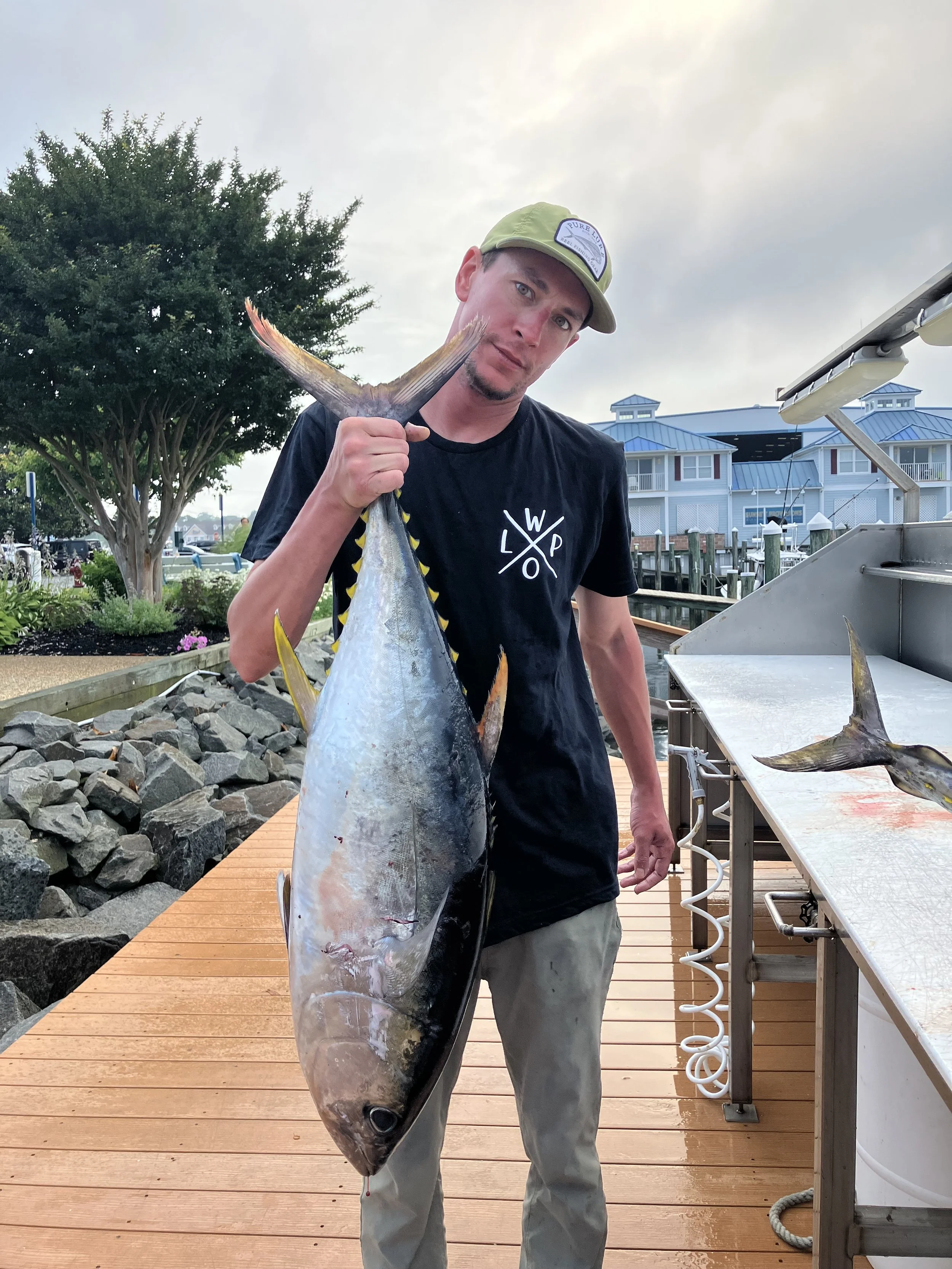 Man holding a large fish on a dock near boats and a waterfront house, overcast sky.
