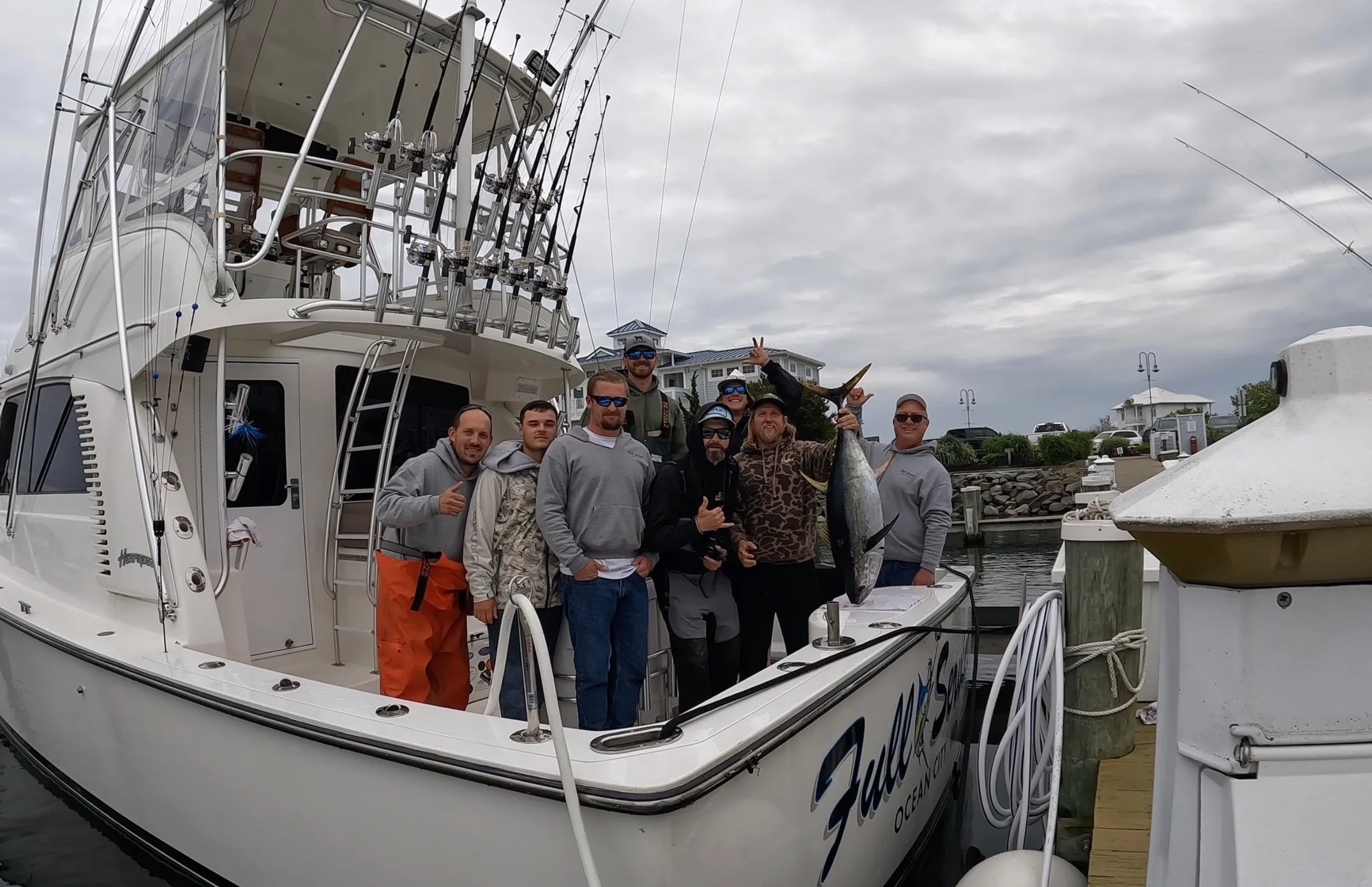 Group of people on a boat holding a large fish, with fishing rods on the boat and a marina in the background.