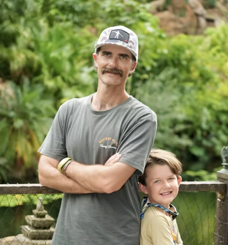 A man and a young boy stand outdoors in front of a wooden fence with lush green foliage in the background. The man has a mustache, wears a cap, and a gray T-shirt with his arms crossed. The boy is smiling and has short brown hair, wearing a tan shirt with a camera strap around his neck.