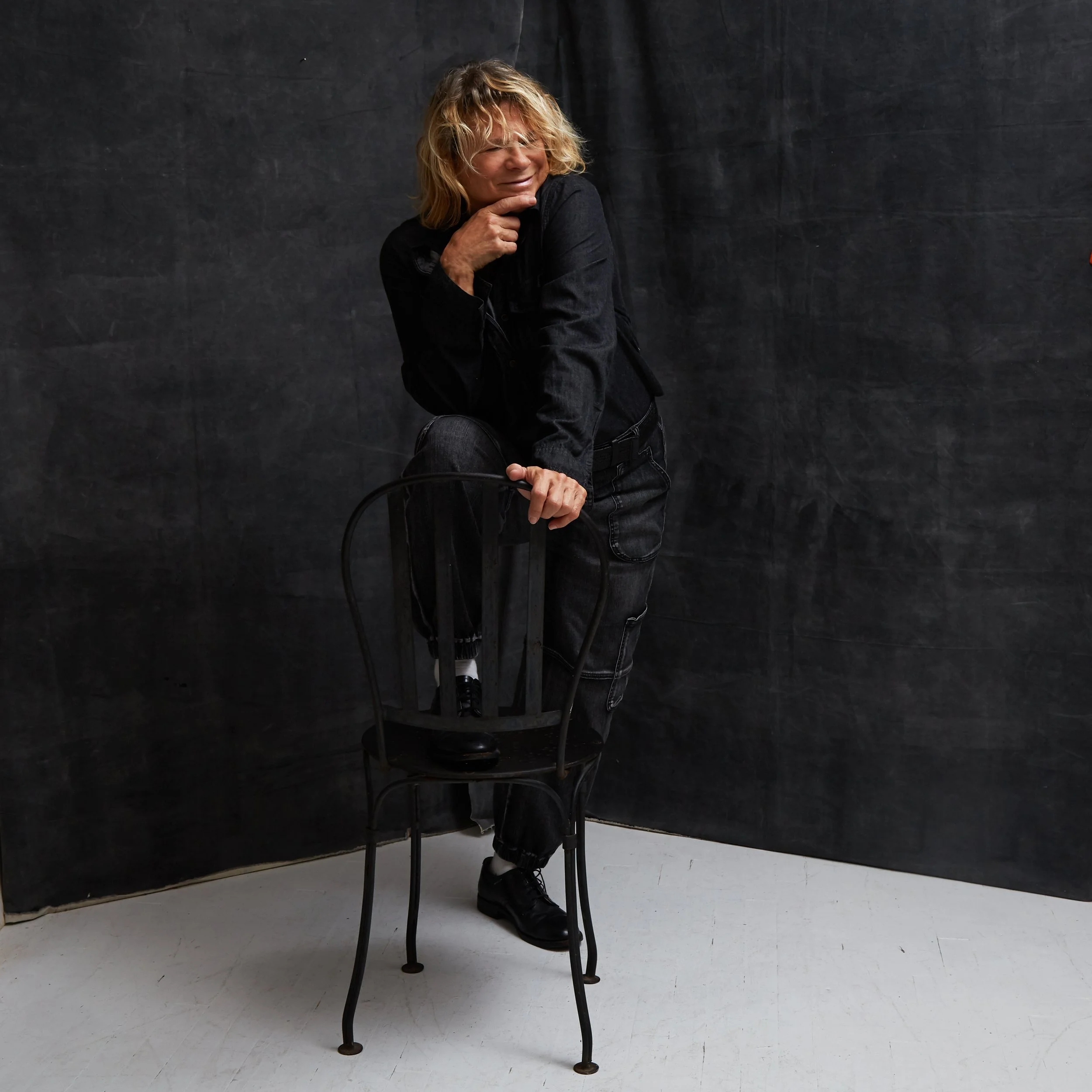Woman with blonde curly hair sitting on a black wooden chair against a dark wall, smiling with her hand on her chin, wearing a black jacket and jeans.