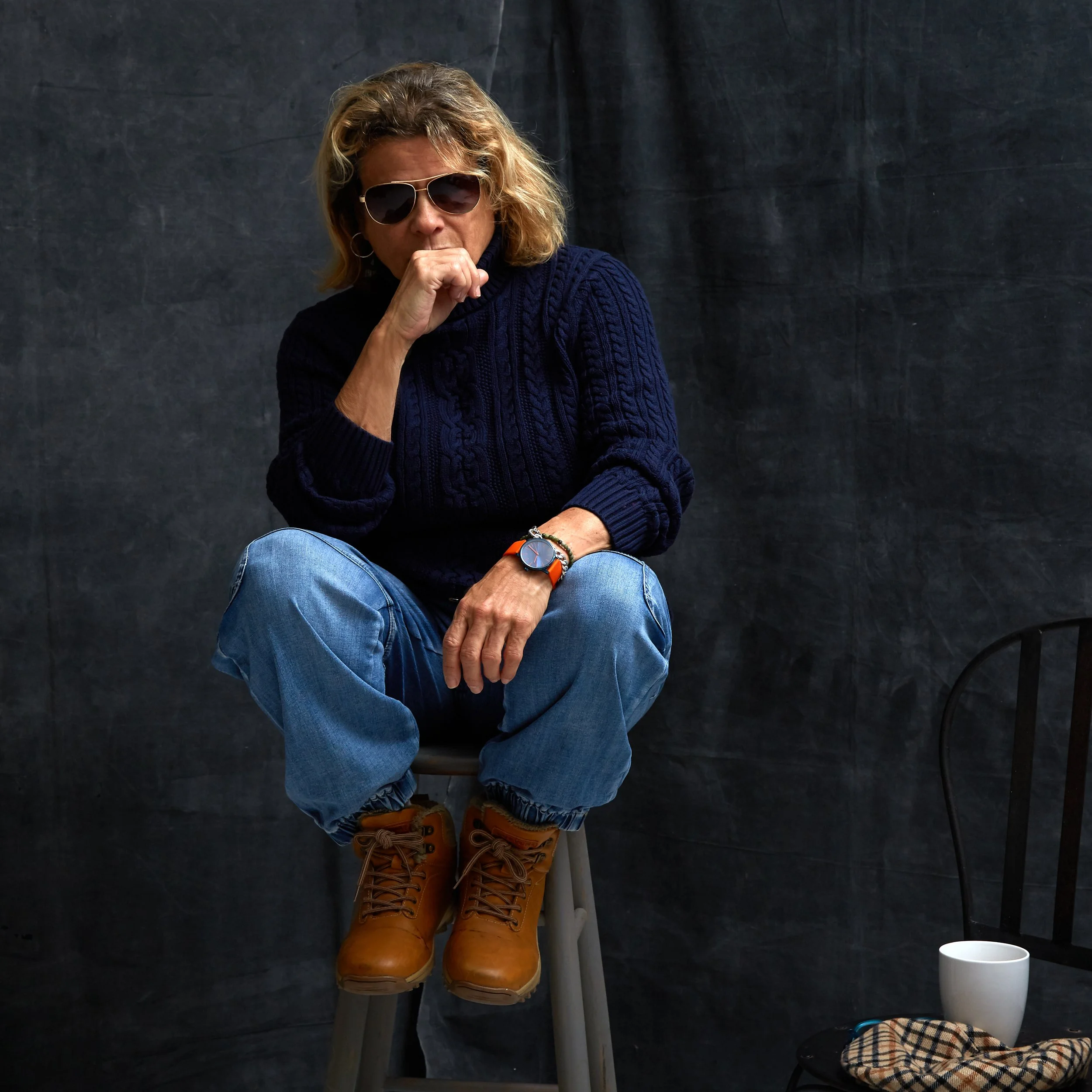 A woman with blonde hair, wearing sunglasses, a navy cable-knit sweater, and tan hiking boots, sits on a ladder in front of a dark backdrop, with a white mug and a plaid cloth on the table nearby.