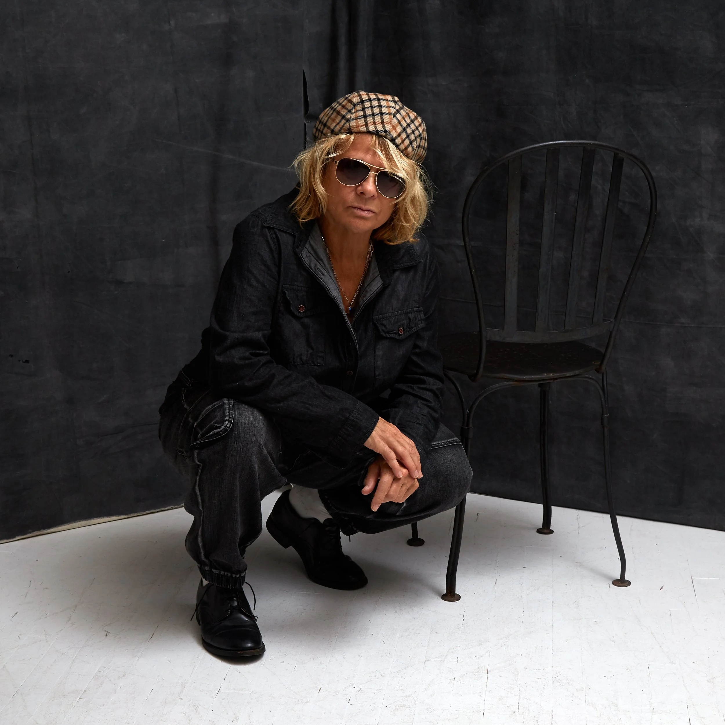 A woman with blonde wavy hair, wearing sunglasses, a plaid beret, a black denim jacket, and black boots, sitting on a black chair against a dark gray background.
