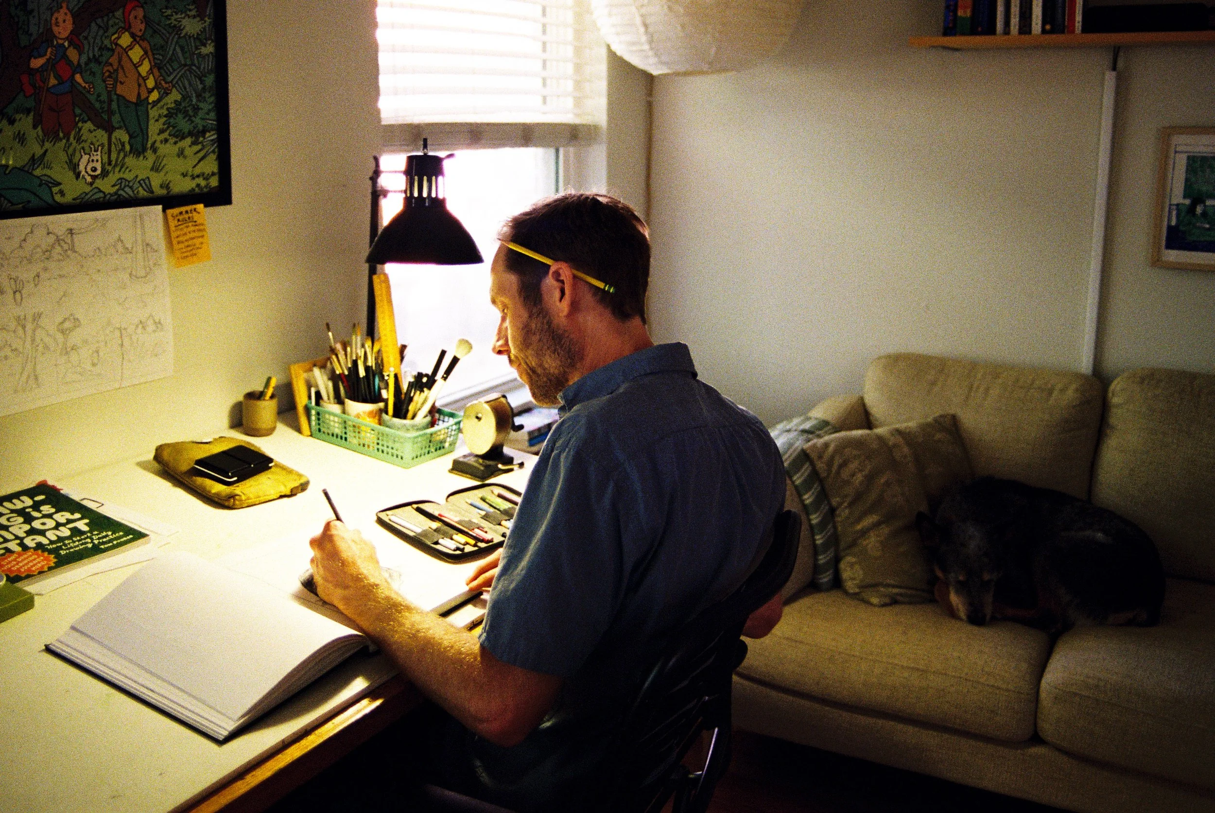 A man sitting at a desk, working on a sketch or drawing with art supplies, in a cozy room with a bookshelf and couch in the background.