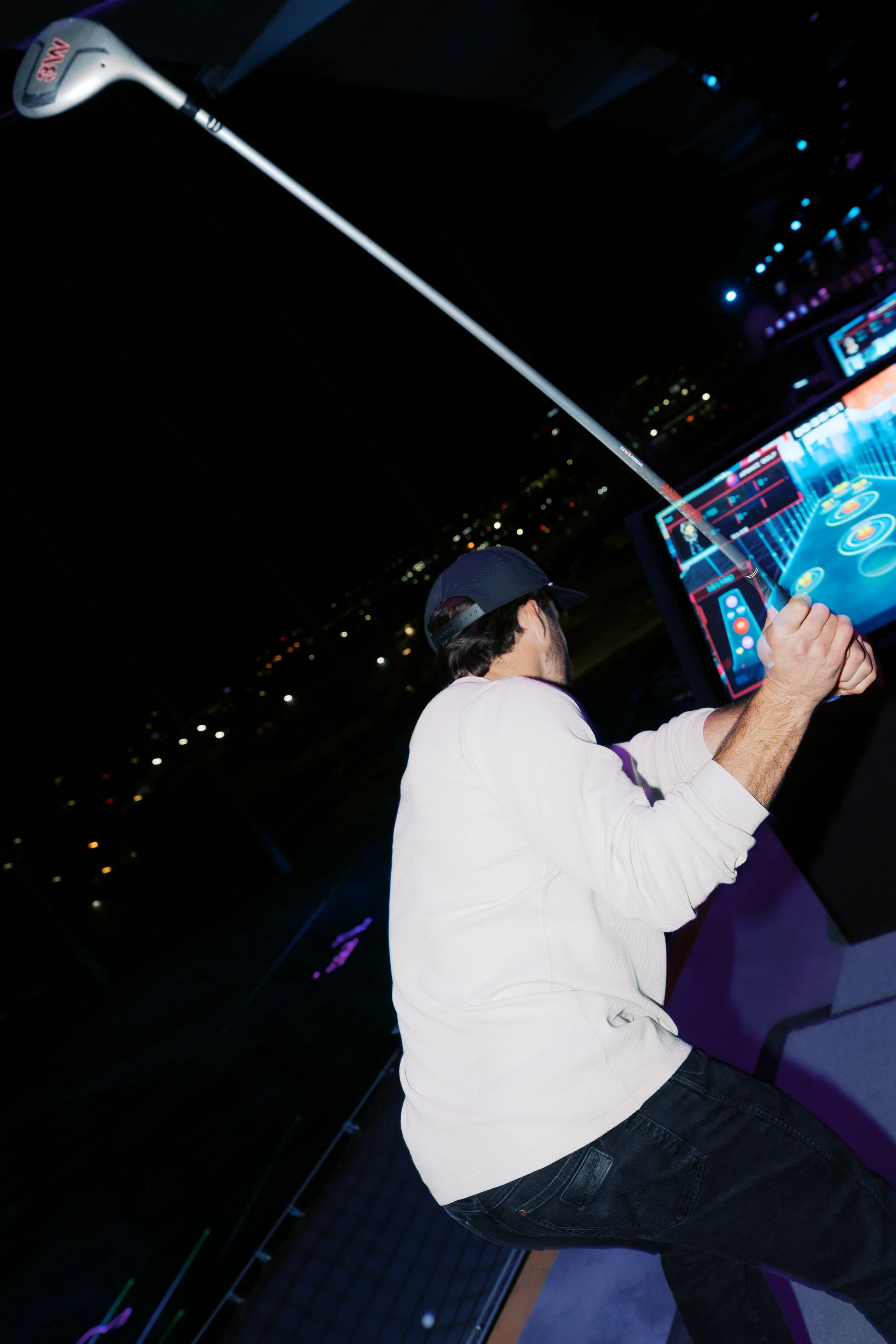 A man in a white sweatshirt and black jeans playing mini golf at night, holding a golf club with a cityscape in the background.