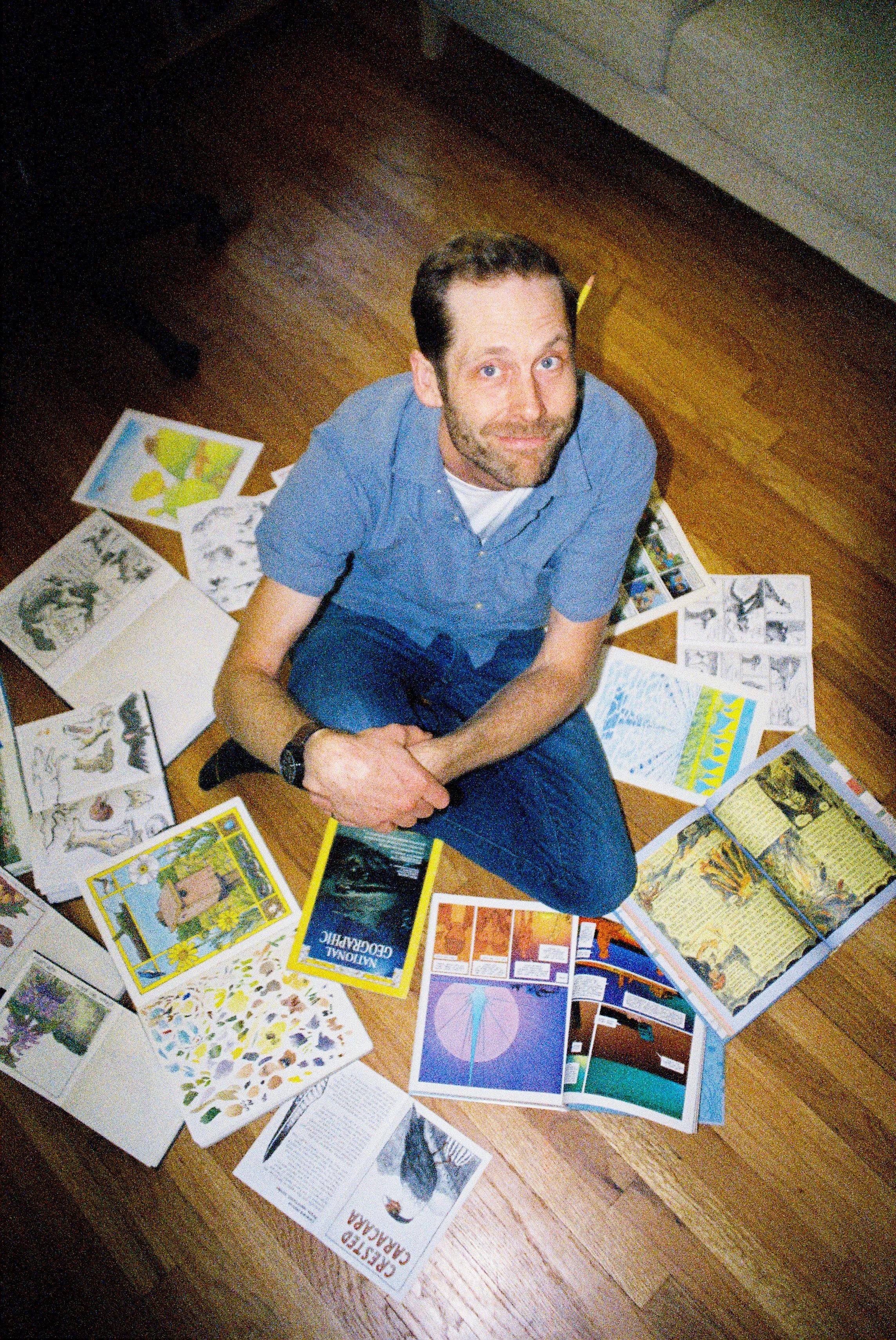 A man with brown hair and a beard, sitting on the floor surrounded by various books and educational materials, looking up at the camera.