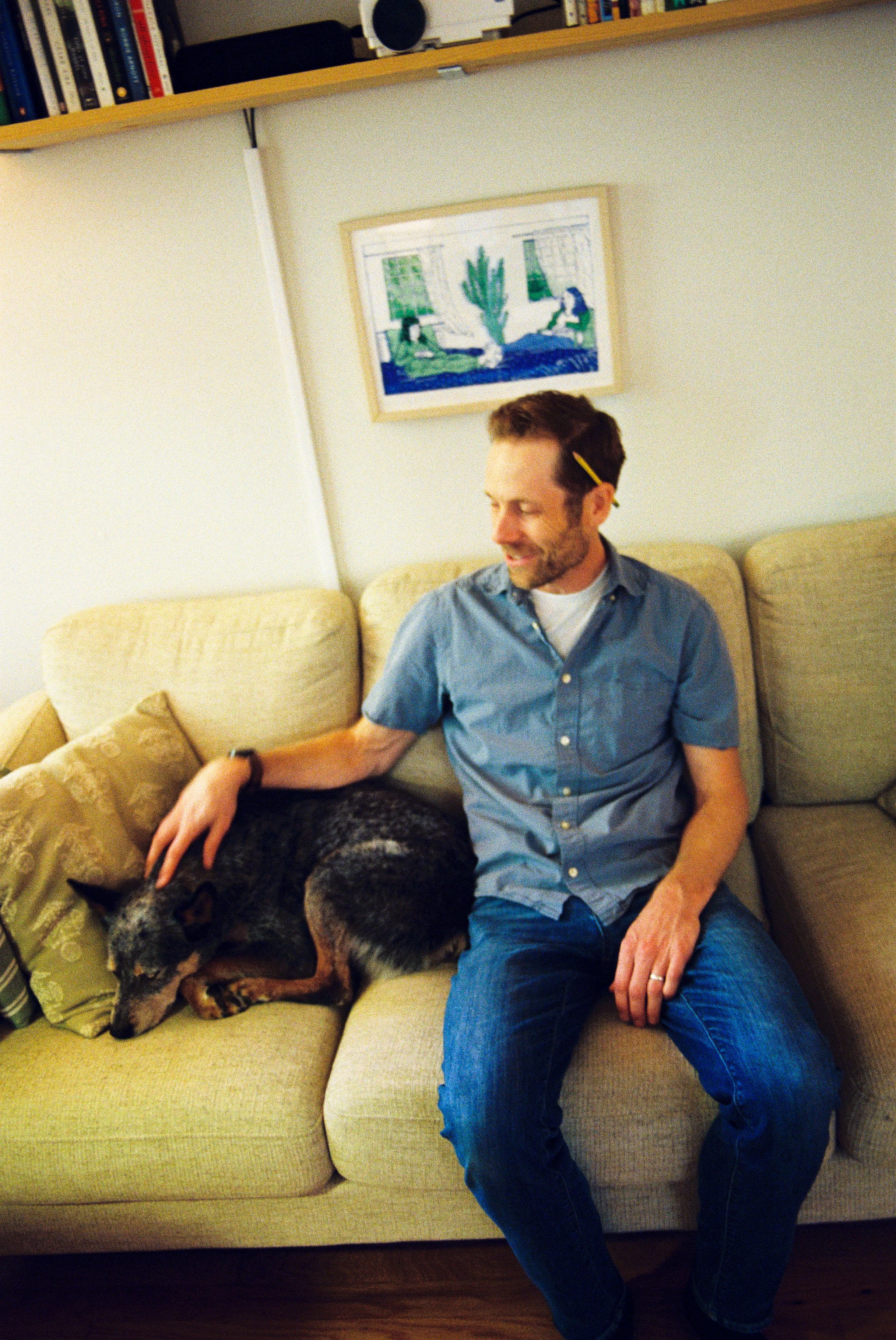 A man sitting on a beige couch with a large dog resting beside him. The man is smiling, wearing a blue shirt and jeans. The dog is curled up with its head on a pillow. There is a bookshelf with books above them and a framed artwork on the wall behind