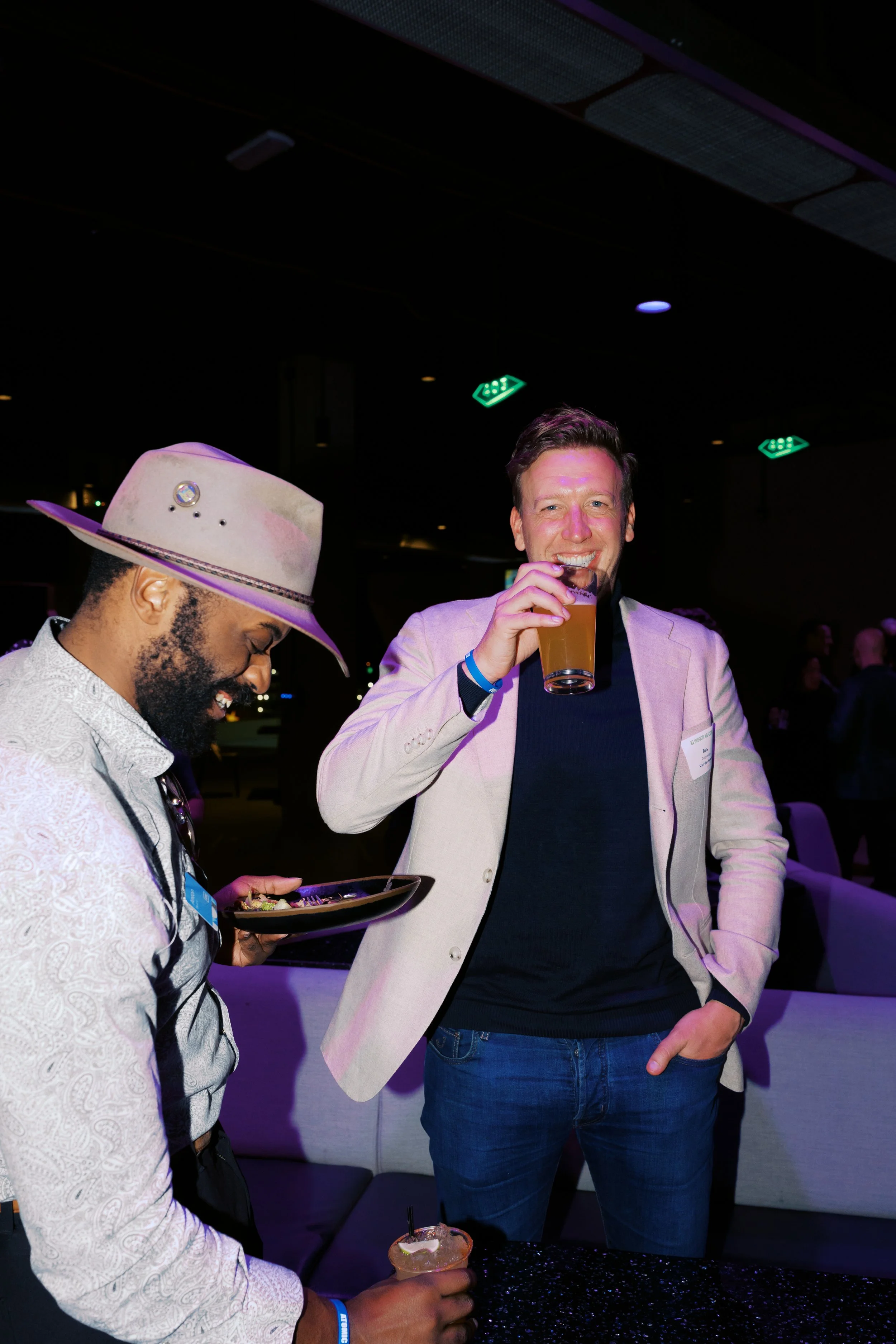 Two men at a social event, one with a hat and the other in a blazer, enjoying drinks and chatting in a dimly lit venue with neon signs.