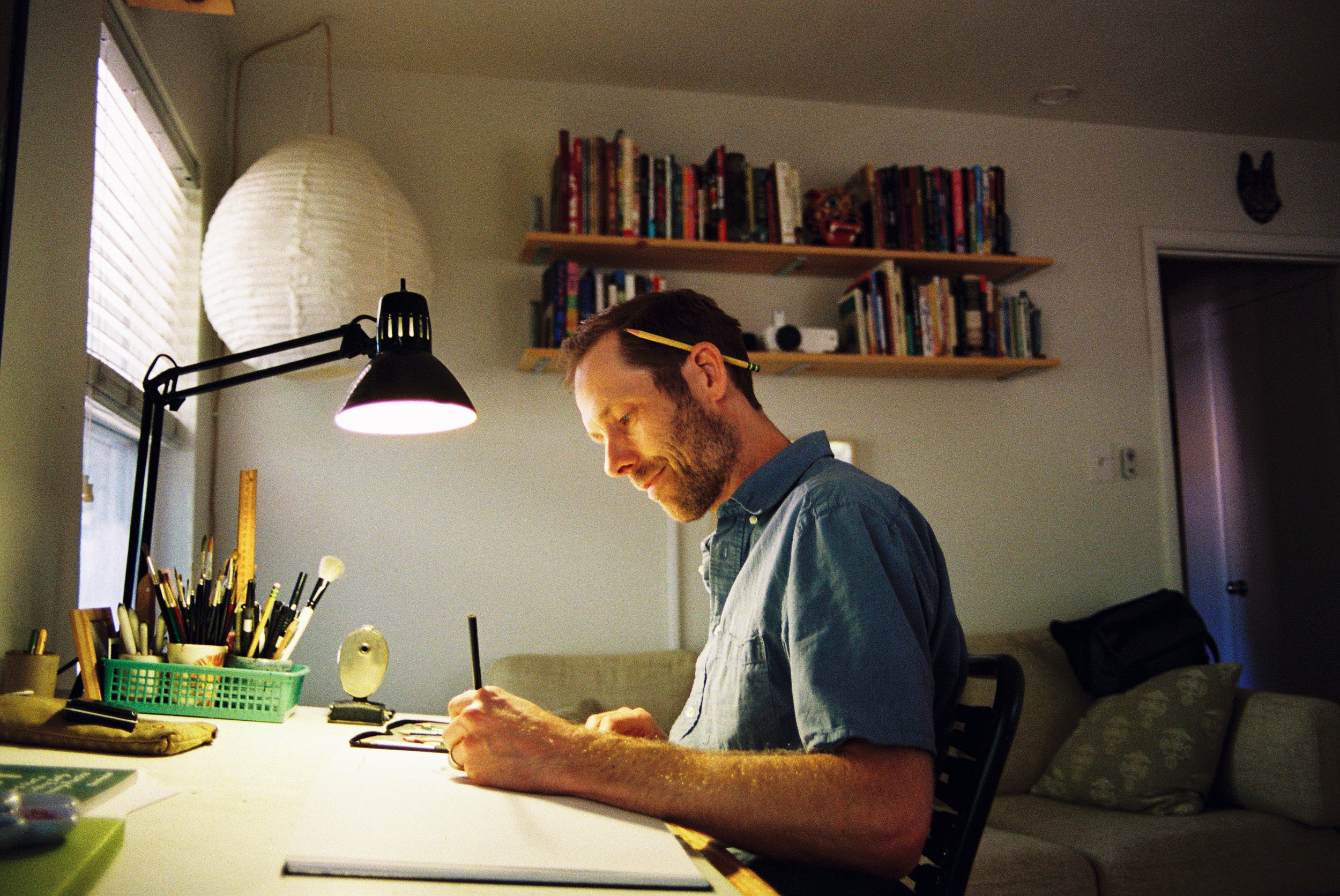 A man with a pencil behind his ear is working on jewelry at a well-lit desk in a room with bookshelves, a large paper lantern, and a window.