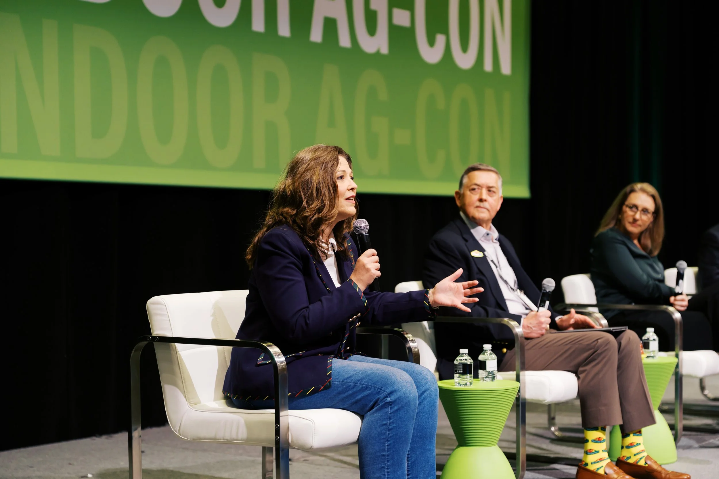 Panel of three people seated on stage at a conference. The woman in the foreground is speaking into a microphone, wearing a navy blazer, jeans, and colorful socks. The man next to her is wearing a suit and observing, while a woman on the far right is wearing glasses and black blazer. Water bottles on tables are visible in front of them.