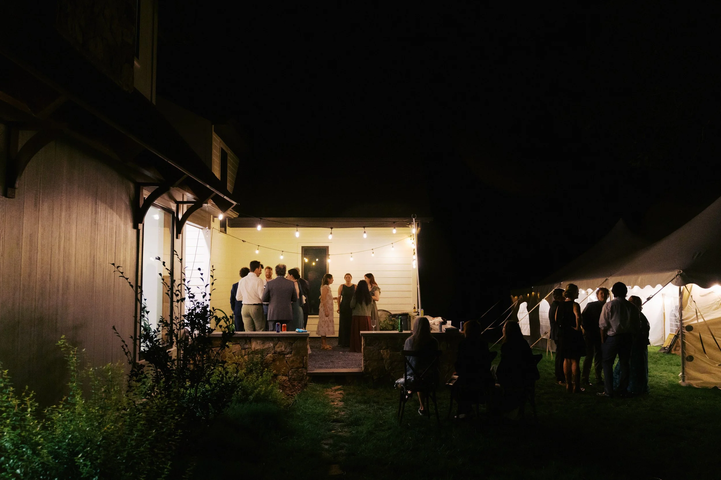 Nighttime intimate wedding reception with people socializing under string lights, featuring a house with a patio, tent, and garden in the background.