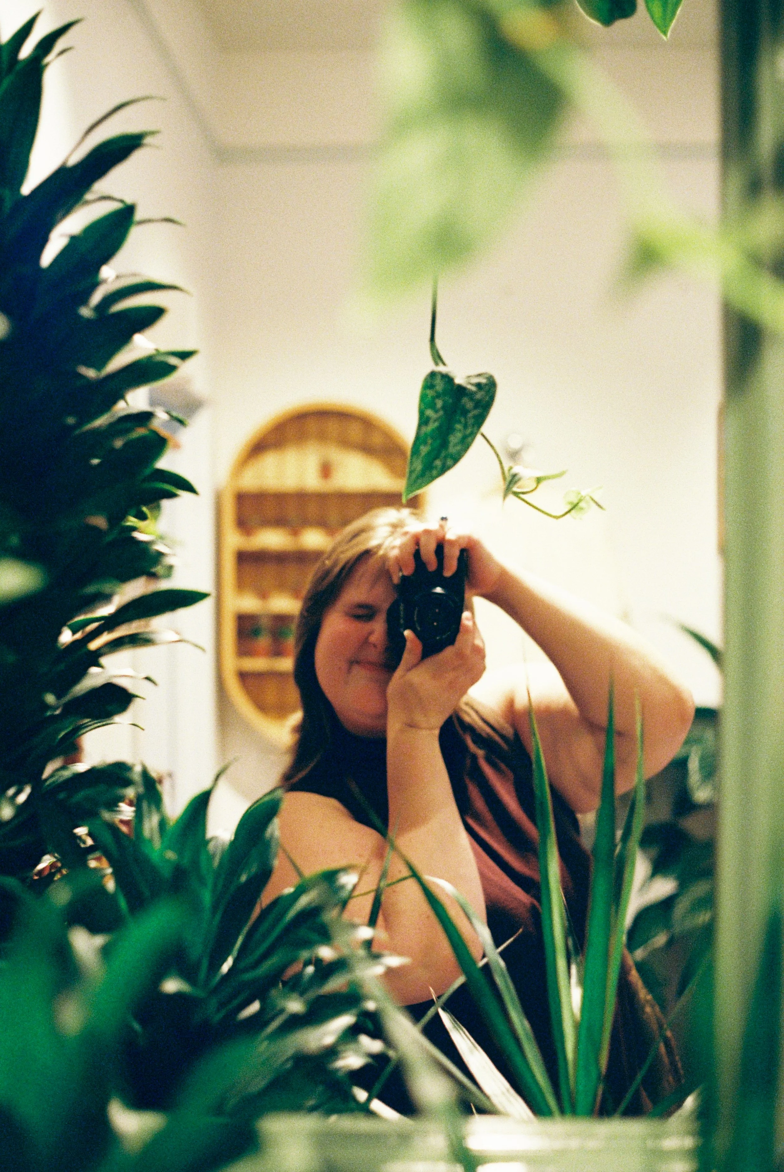 A person taking a selfie with a camera amidst tropical green plants, with their face partially visible and surrounded by large plant leaves.