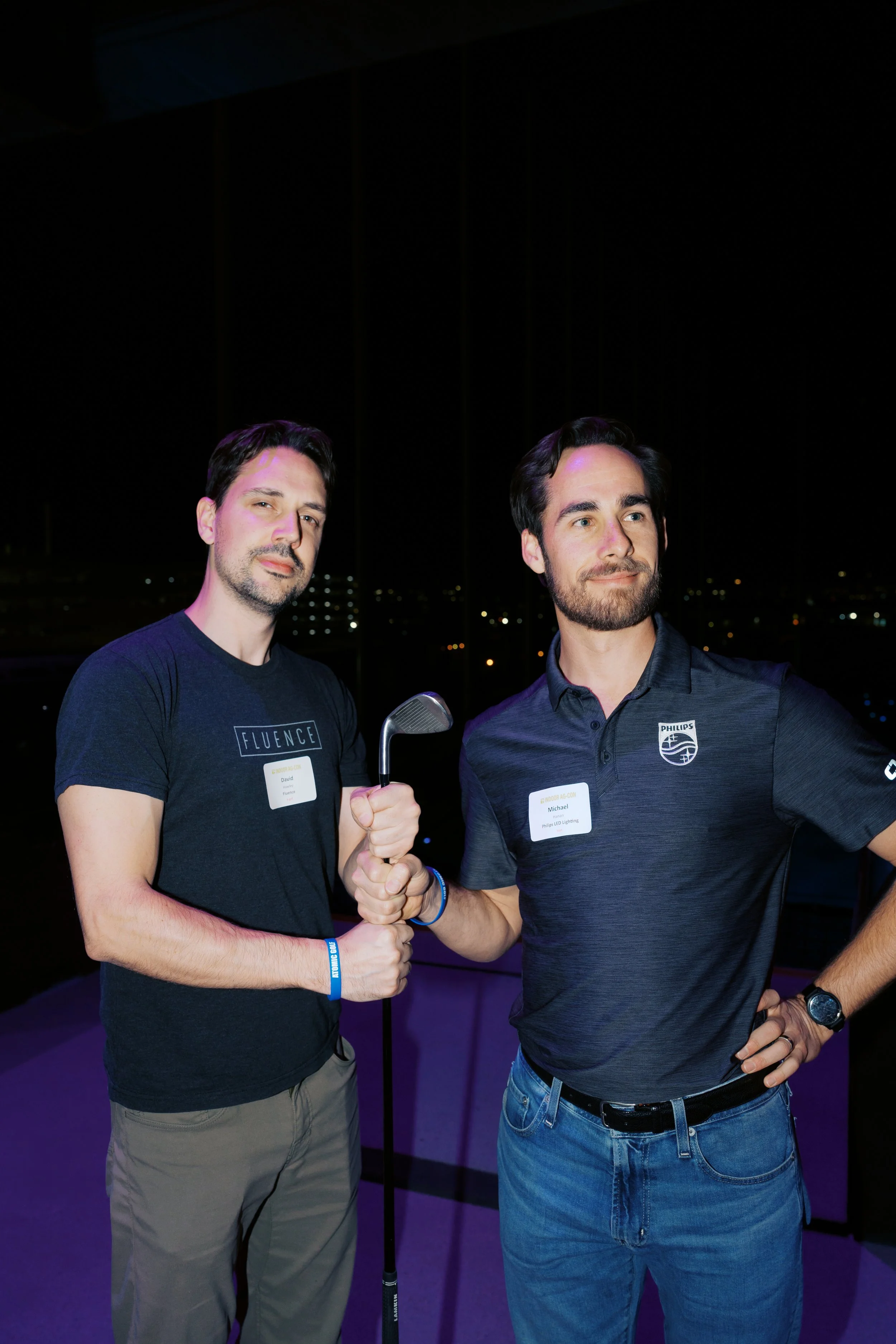 Two men shaking hands at an indoor event, both wearing black shirts with company logos and name tags, with city lights visible outside at night.