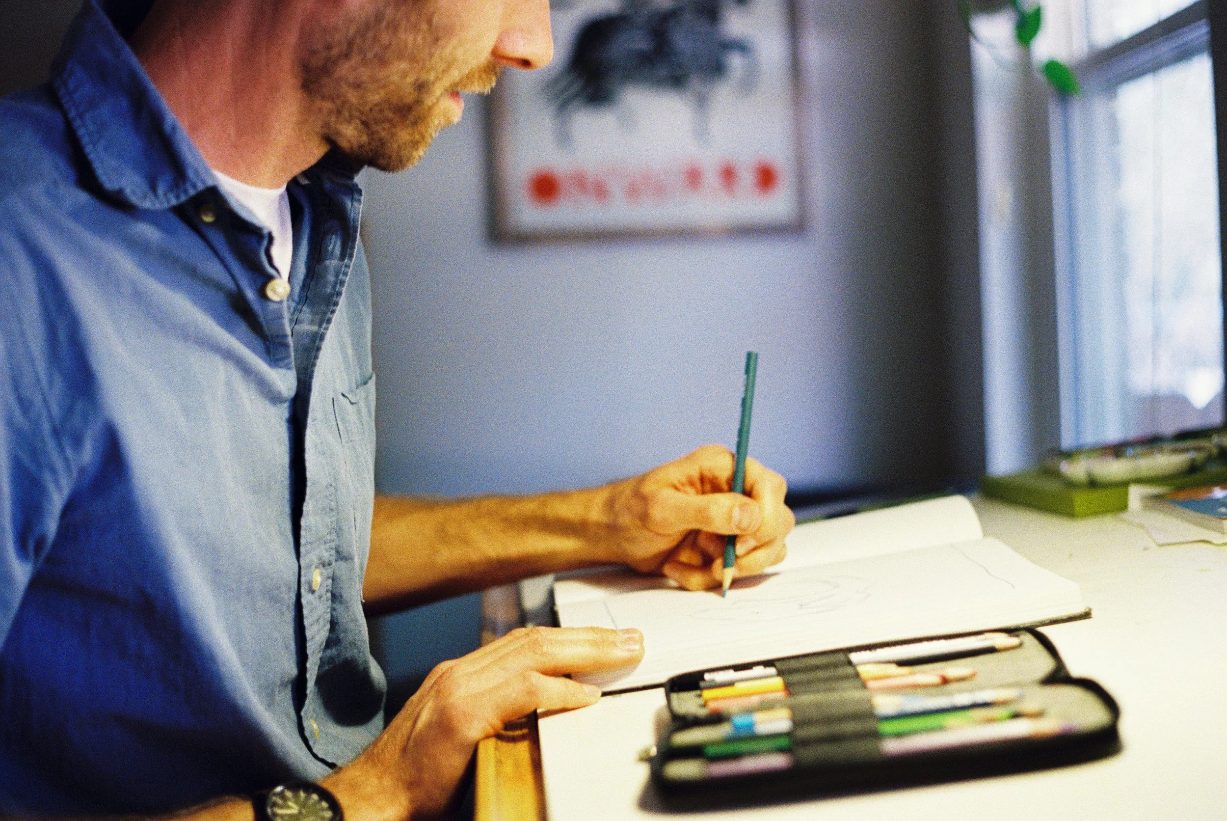 A person wearing a blue shirt is drawing in a sketchbook while sitting at a desk. On the desk, there is a box of colored pencils or markers. In the background, a blurred poster or artwork can be seen on the wall, and a window with a curtain is on the