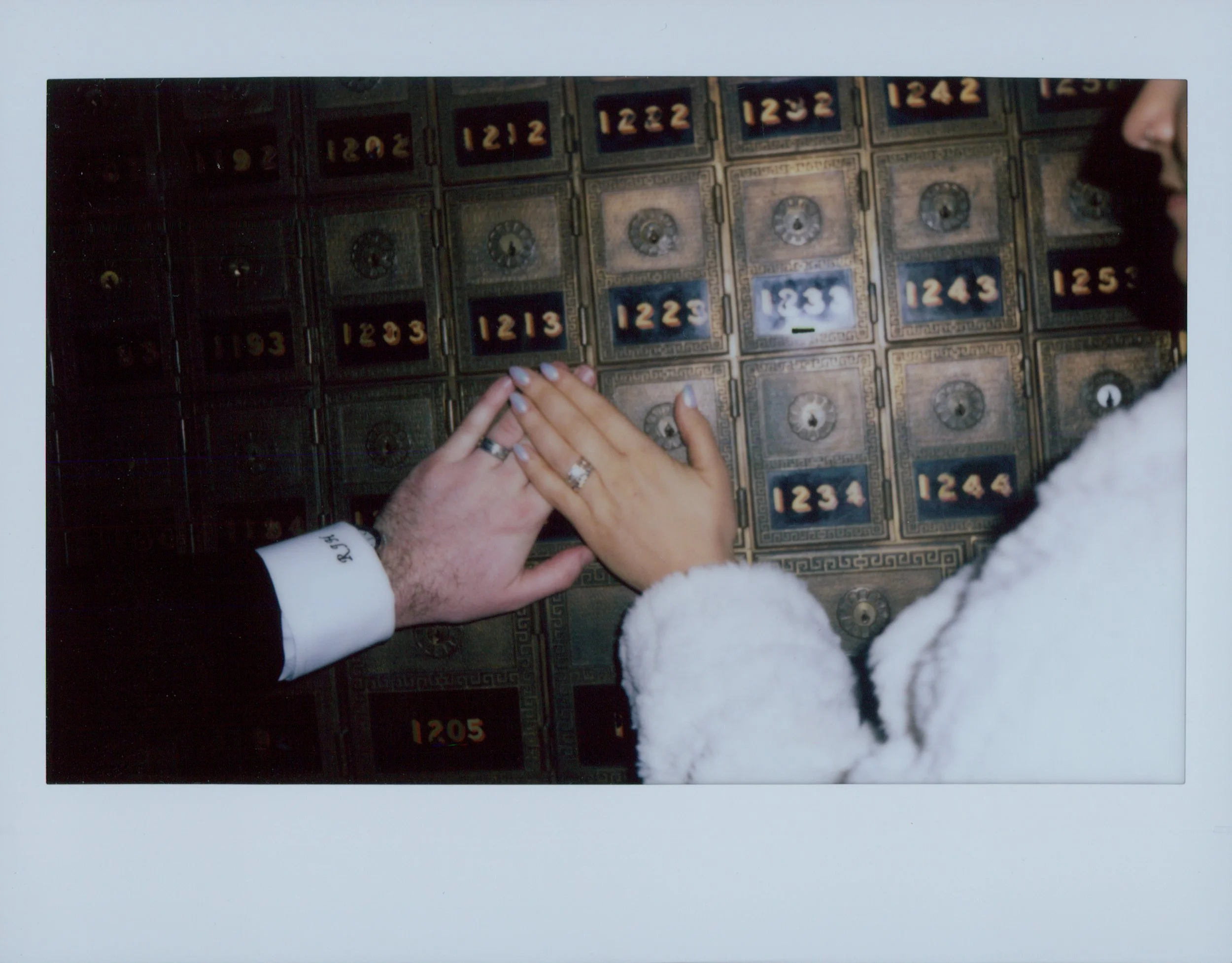 Close-up of multiple vintage metal safes with lock mechanisms and numbered labels, person with light skin and rings on fingers is reaching to open one, partially visible face on right side, background is white.