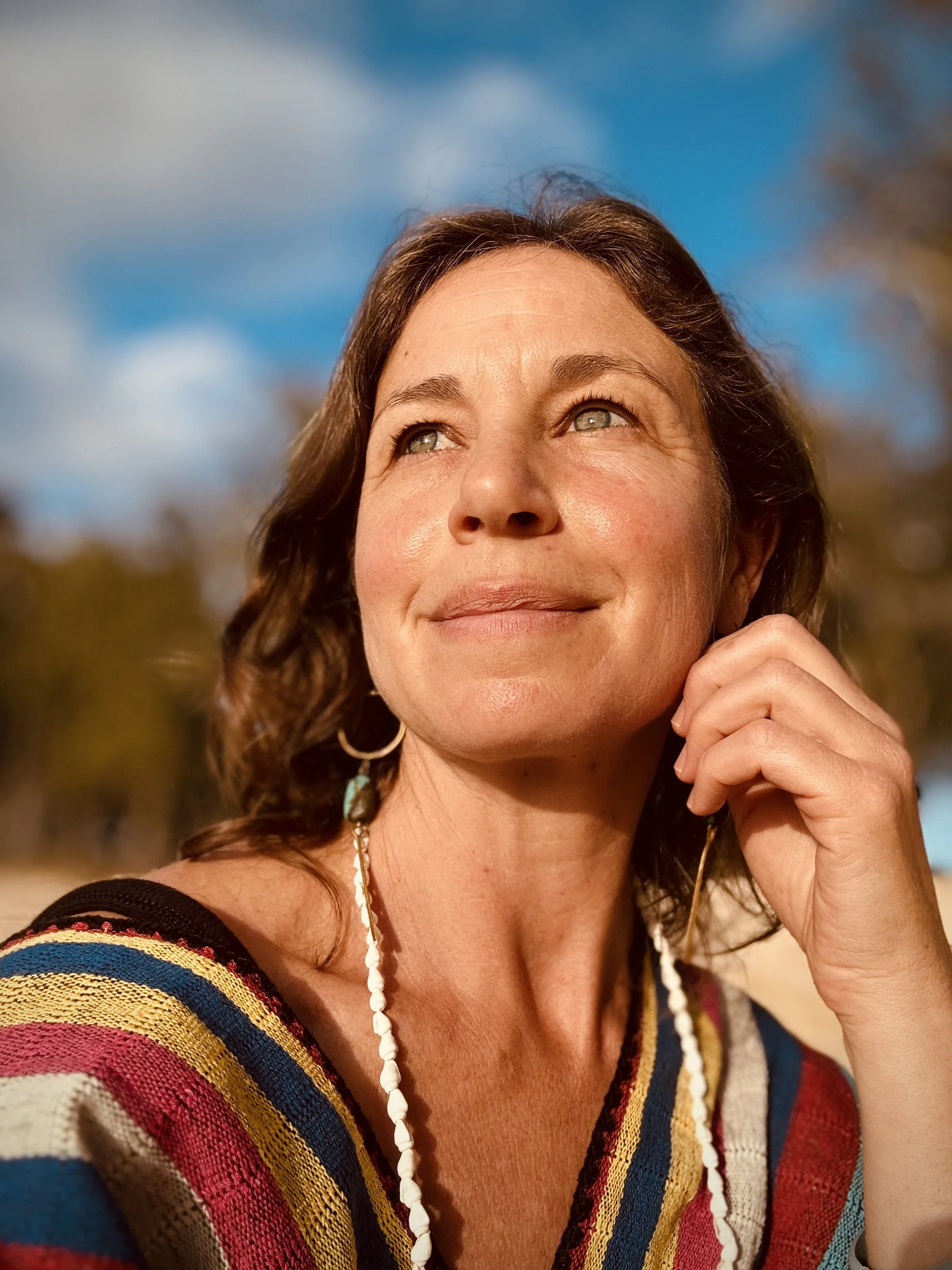 A woman smiling outdoors with a blue sky and blurred trees in the background.