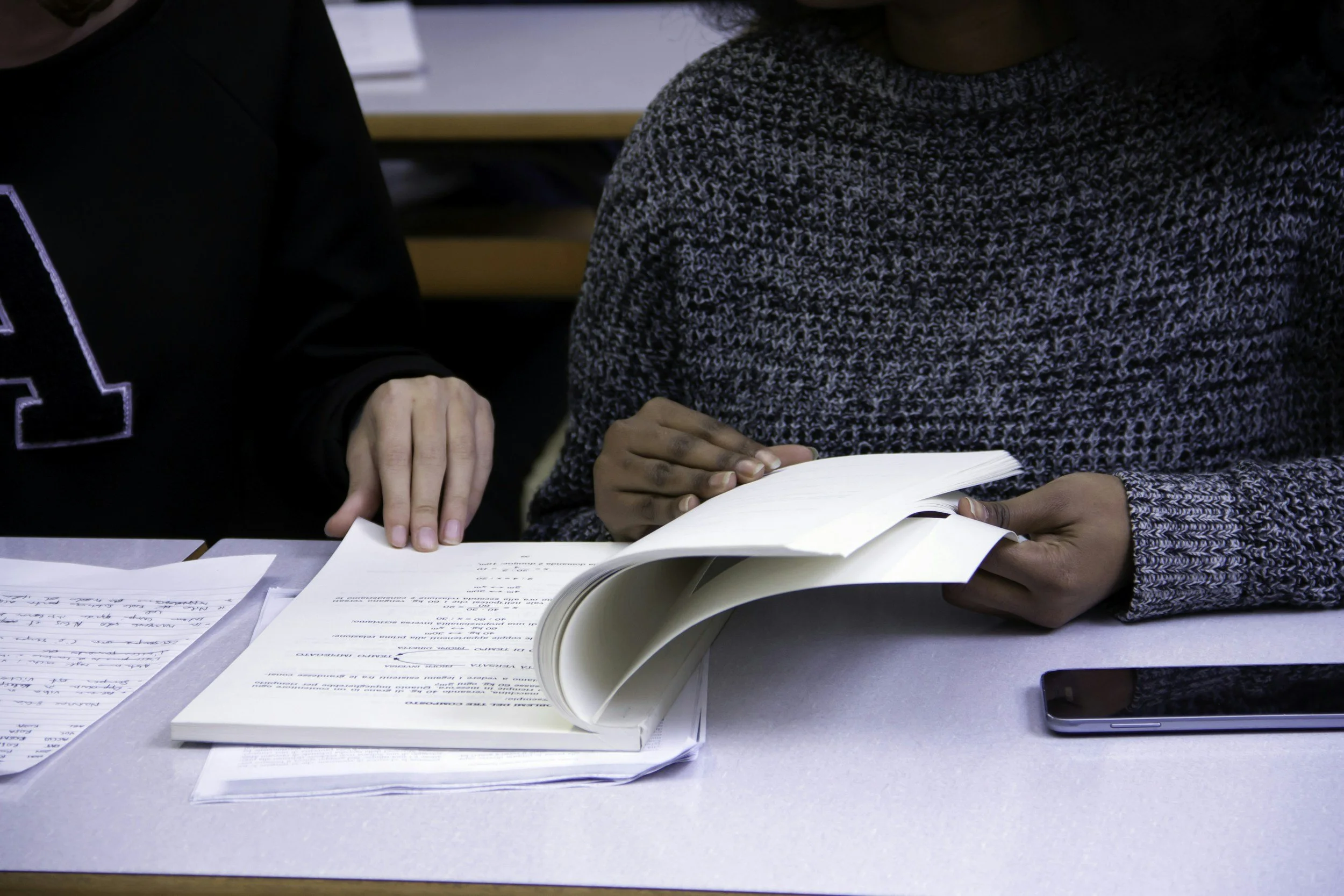 Close-up of a table with scattered papers, a person in a patterned sweater reading a book, and another person pointing at the book.