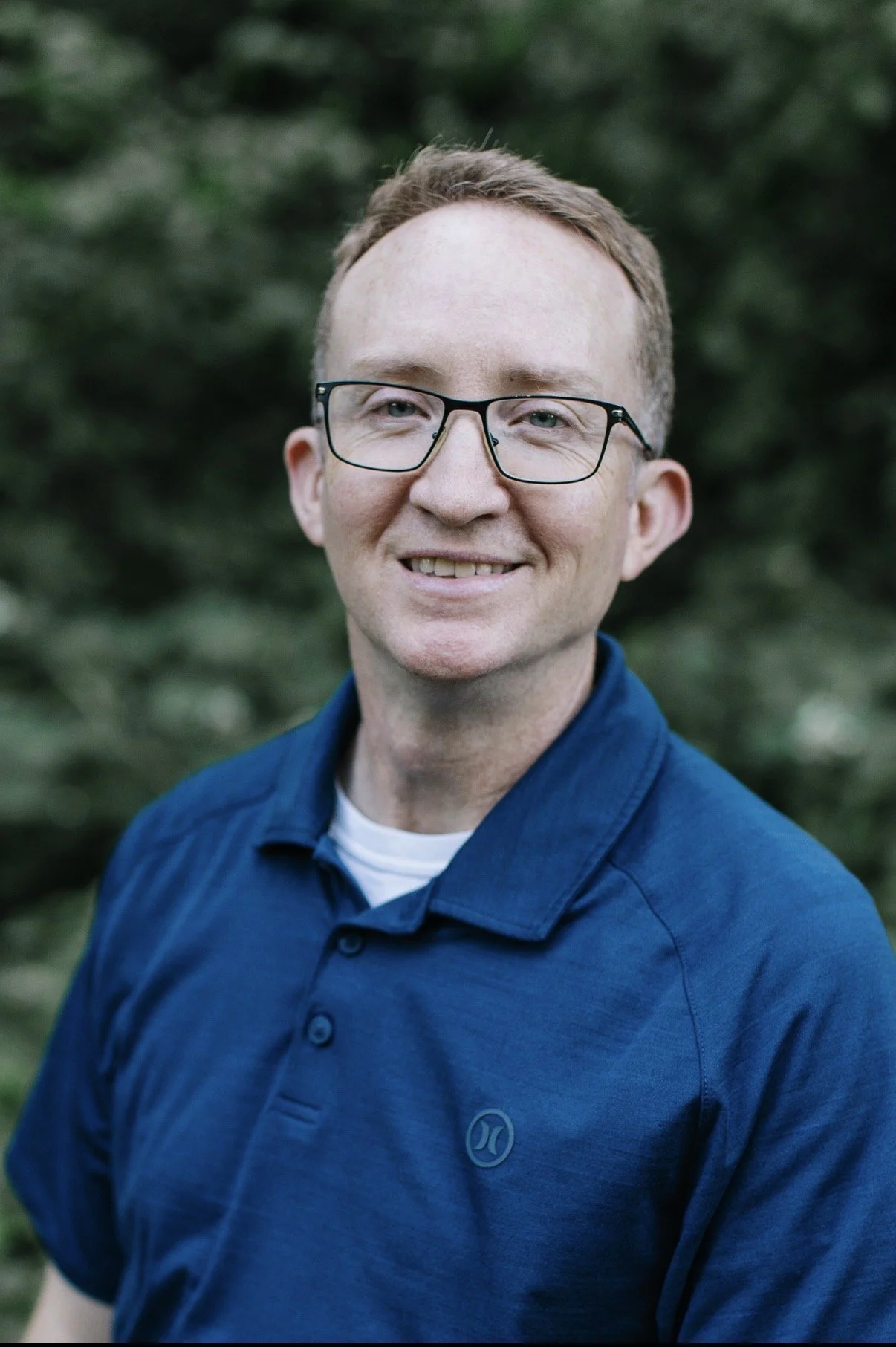 Headshot of a man with glasses smiling, wearing a blue collared shirt, with a blurred green outdoor background.