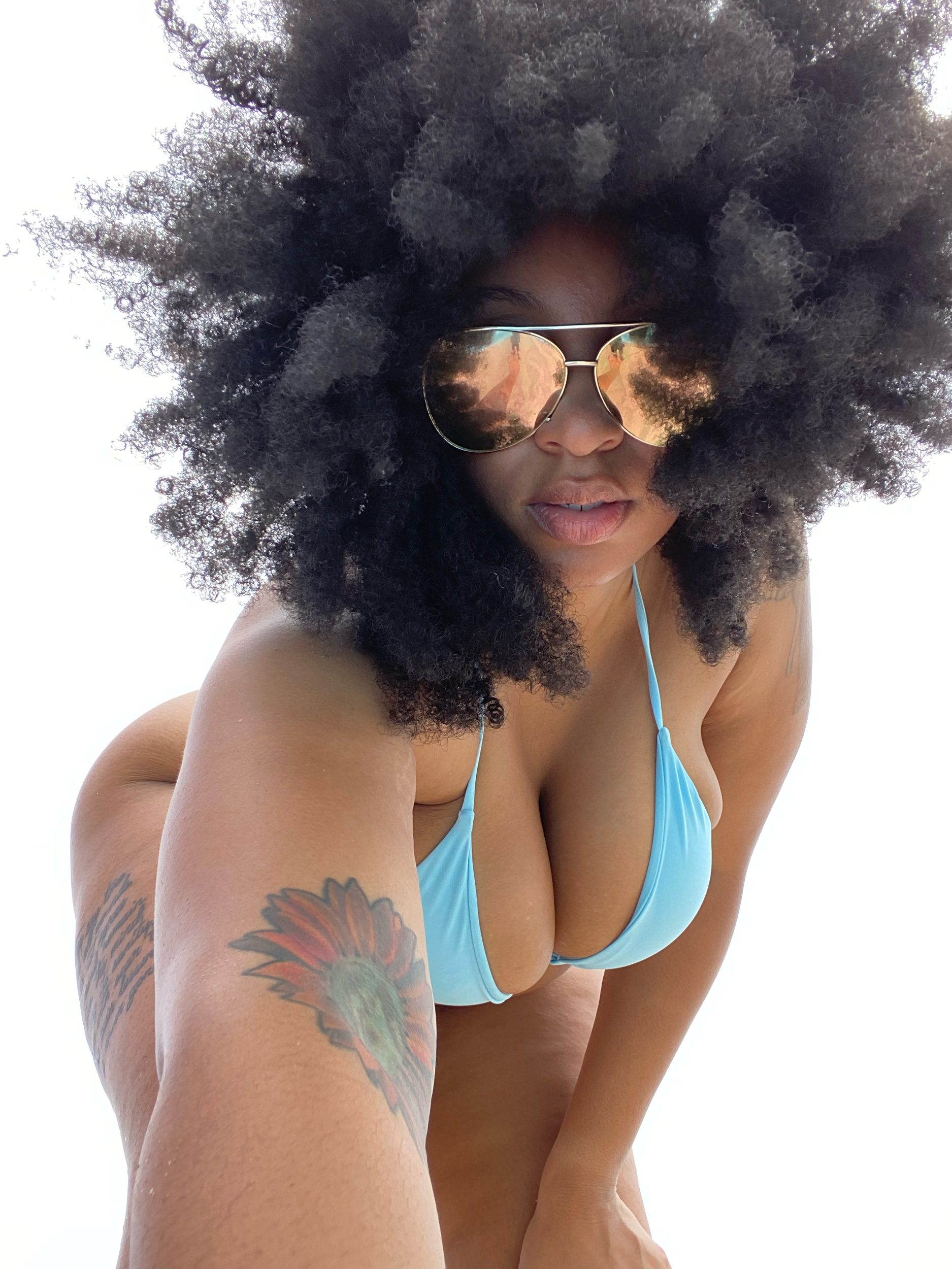 A woman with curly afro hair and sunglasses is wearing a blue bikini top, with visible tattoos on her arm, posing outdoors against a bright sky.