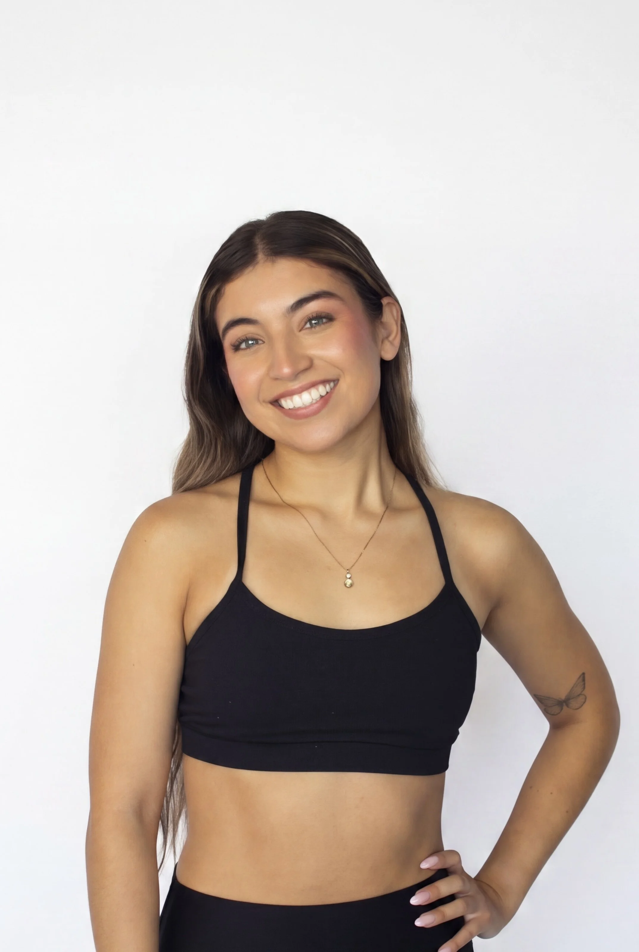 A young woman with long brown hair, blue eyes, and a bright smile, wearing a black sports bra, a delicate gold necklace, and a butterfly tattoo on her left arm, stands against a plain white background.
