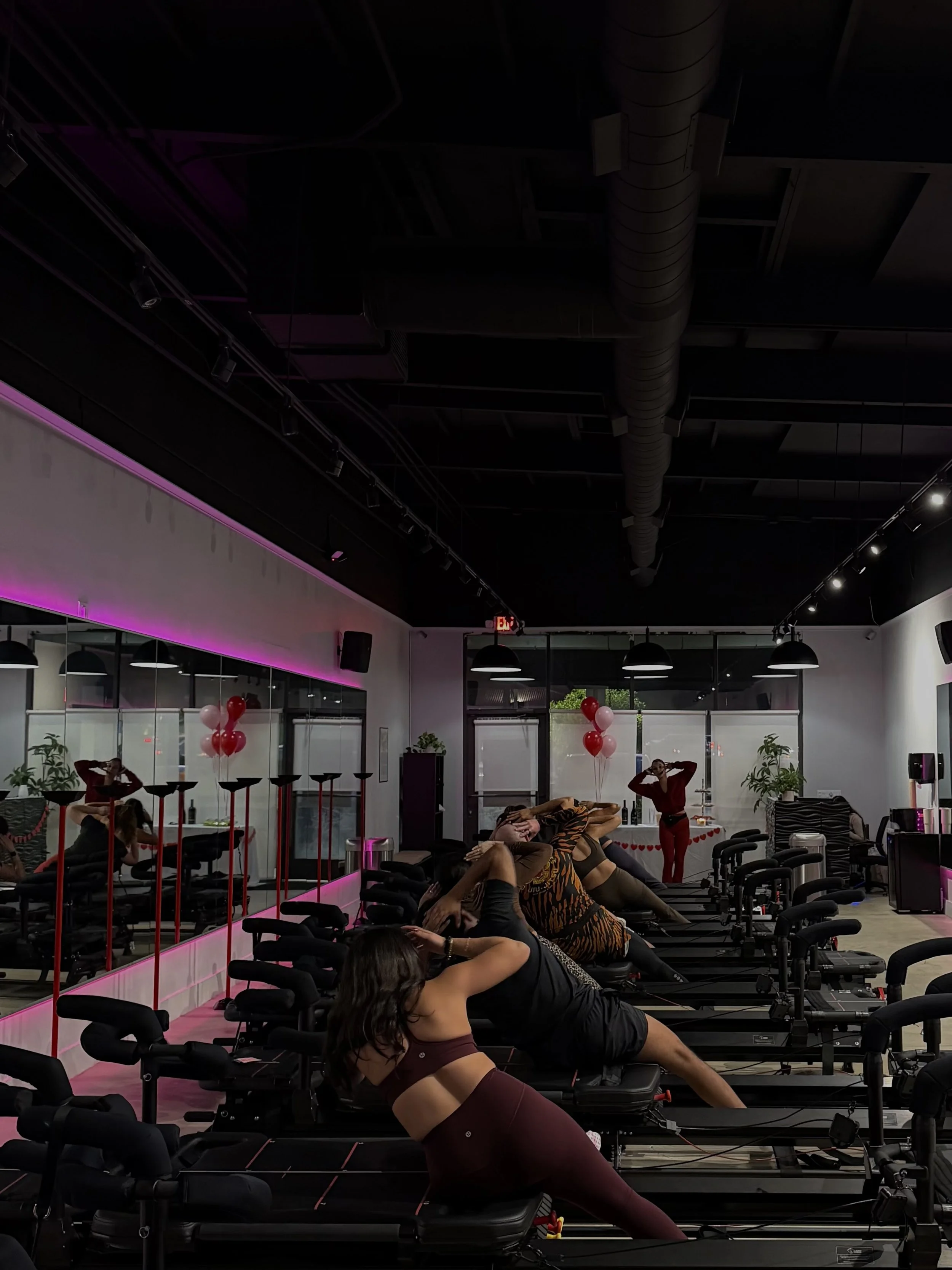 People participating in a lagree fitness class in a lagree fitness studio, lying on megaformer machine in a side plank. The gym has ambient lighting with pink accents, large windows, and ceiling ductwork.