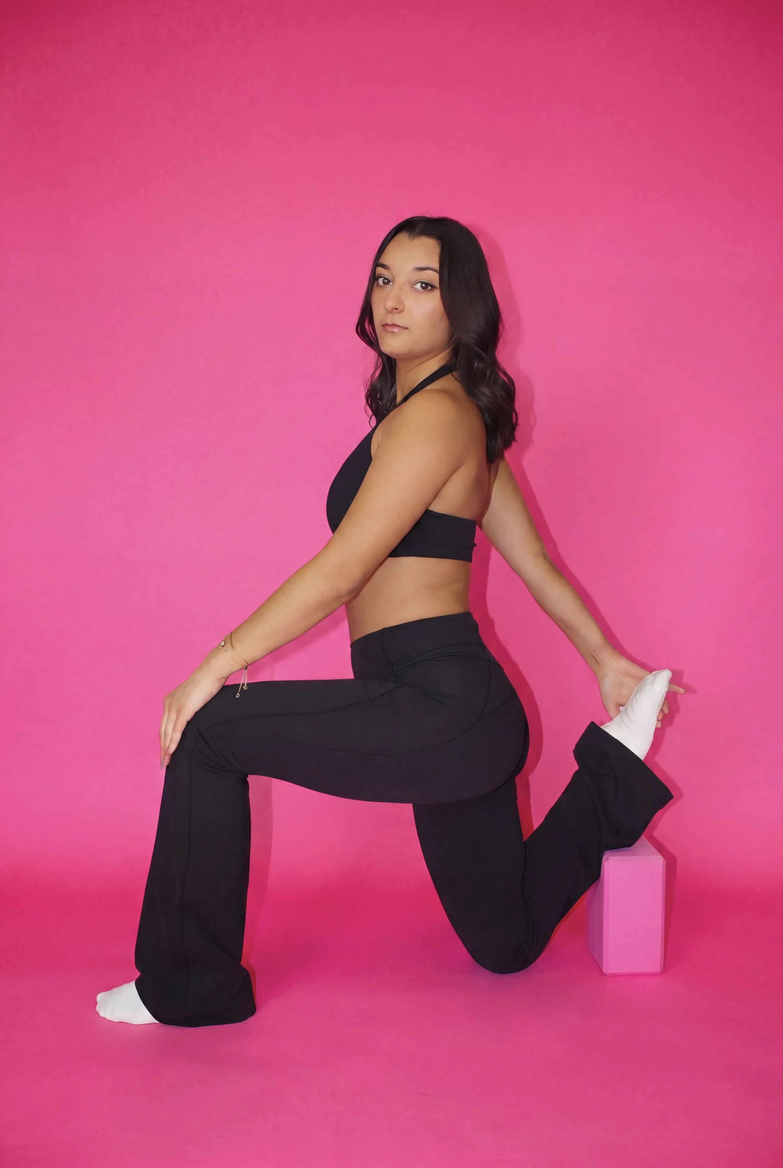 A woman in black activewear performing a yoga pose on a pink background, with one leg bent behind her and held by her hand, using a pink box for support.