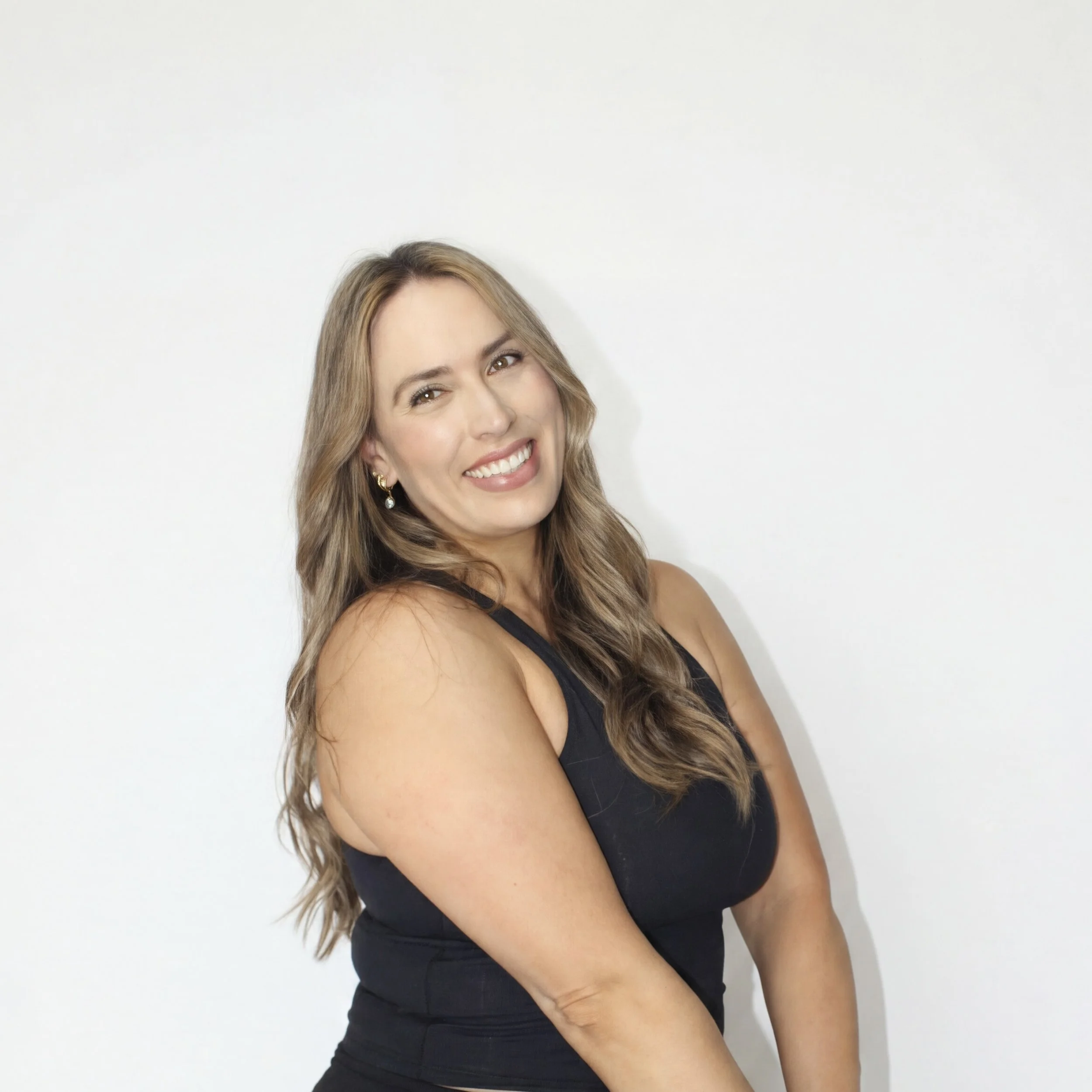 A woman with long, wavy, light brown hair and light skin, smiling and wearing a black sleeveless top, standing against a plain white wall.