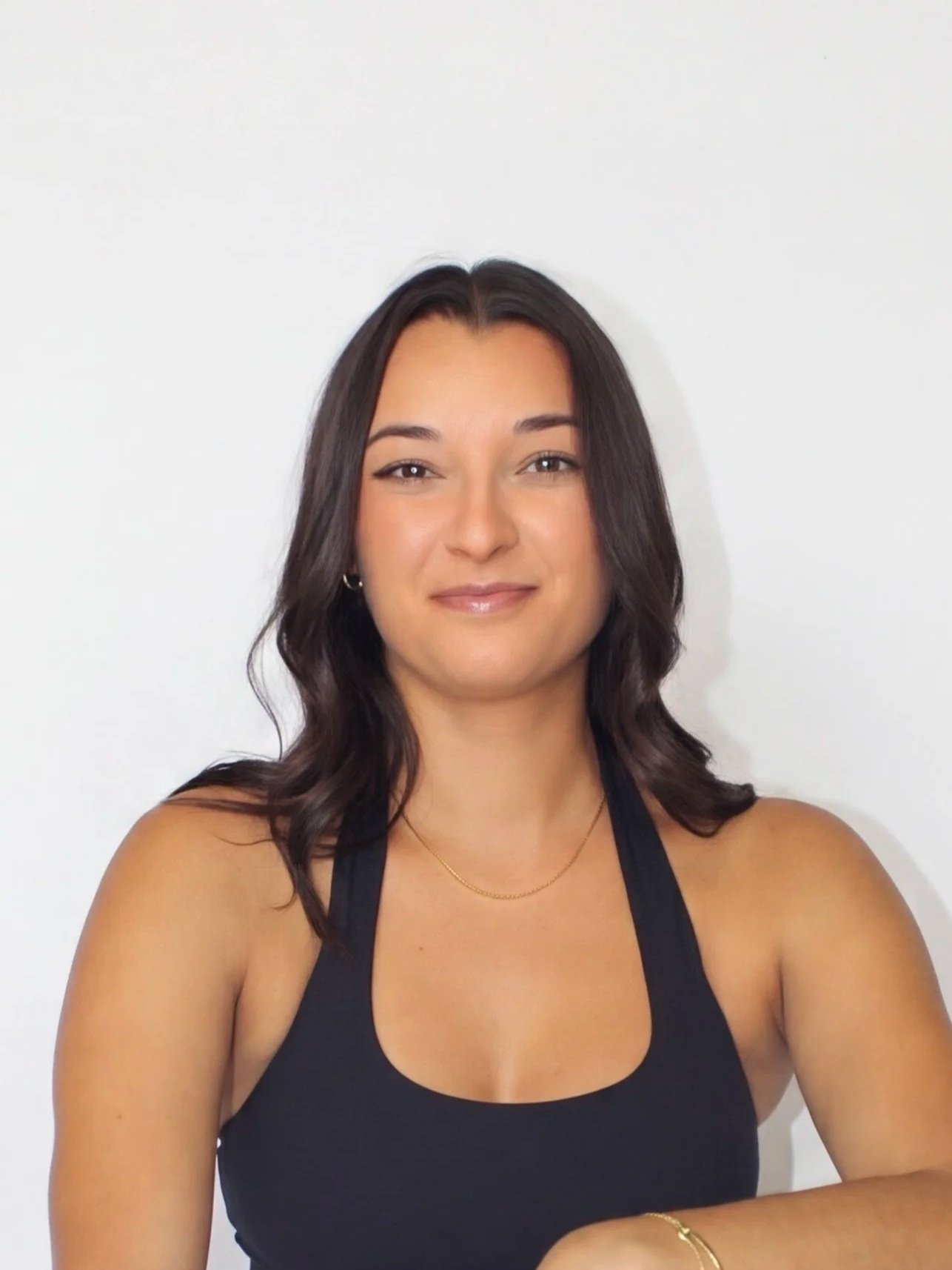 A young woman with long dark hair smiling at the camera, wearing a black tank top and a gold necklace, in front of a plain white wall.