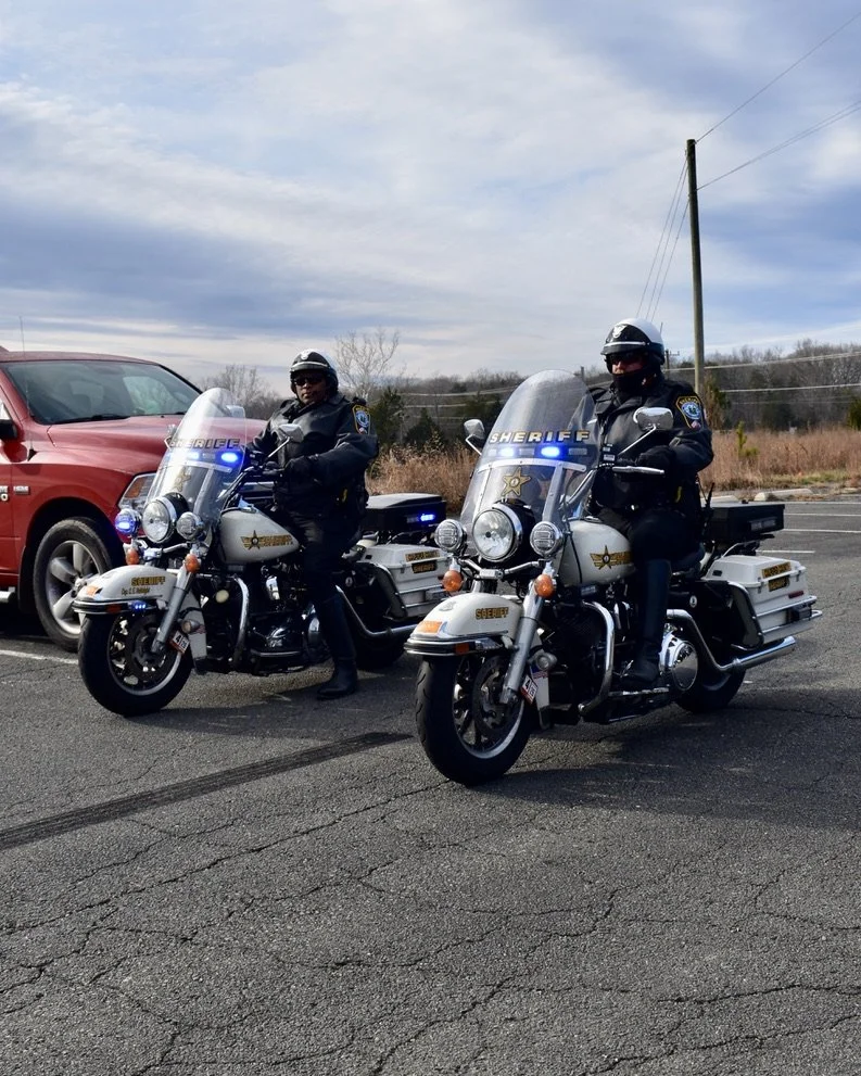 Two sheriff deputies on white patrol motorcycles with blue lights parked in a lot, with a red vehicle and open landscape in the background.