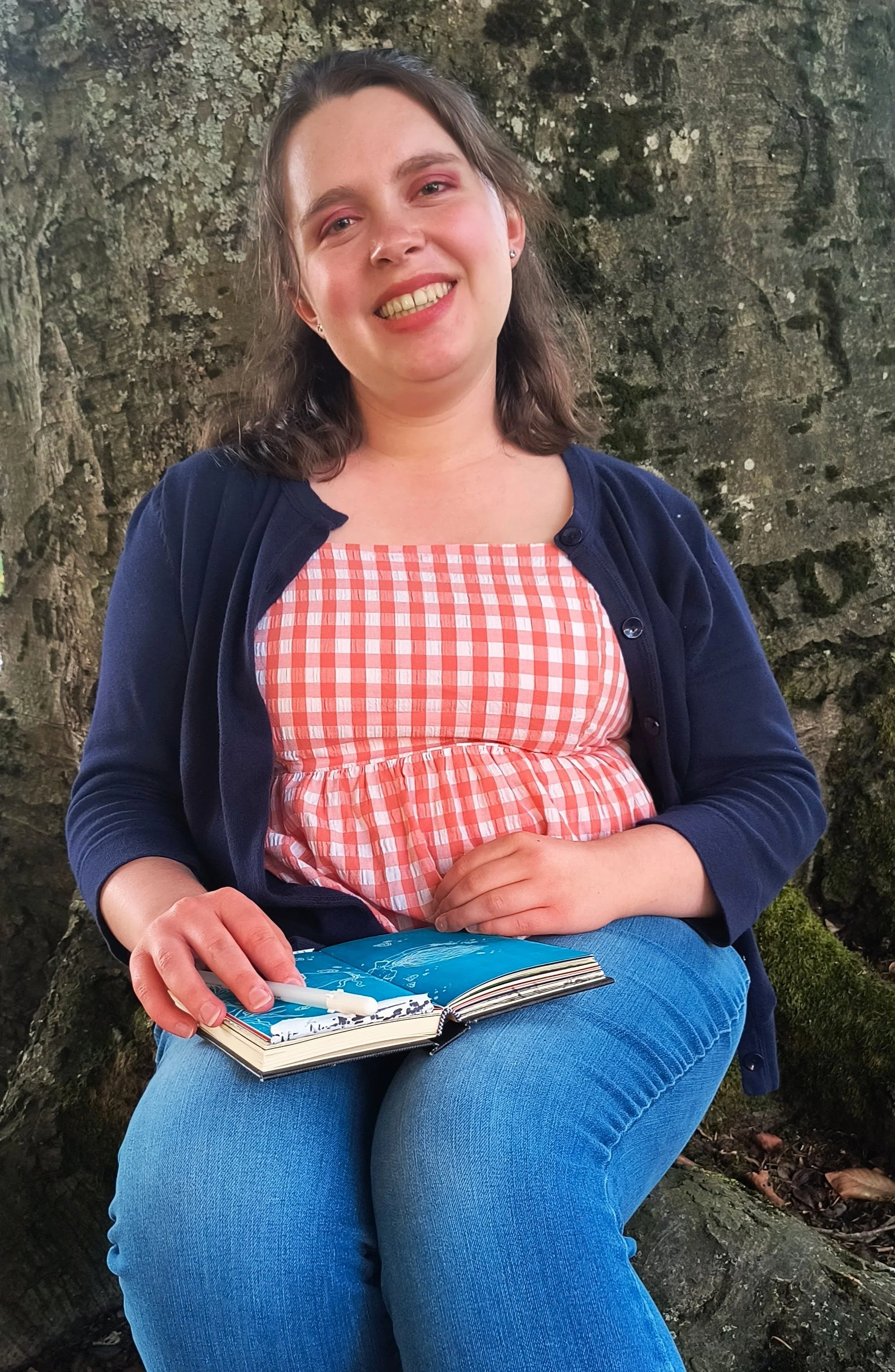 A woman (Ceithlinn Gates) sitting outdoors against a large tree, smiling at the camera, holding a sketchbook and a pen.