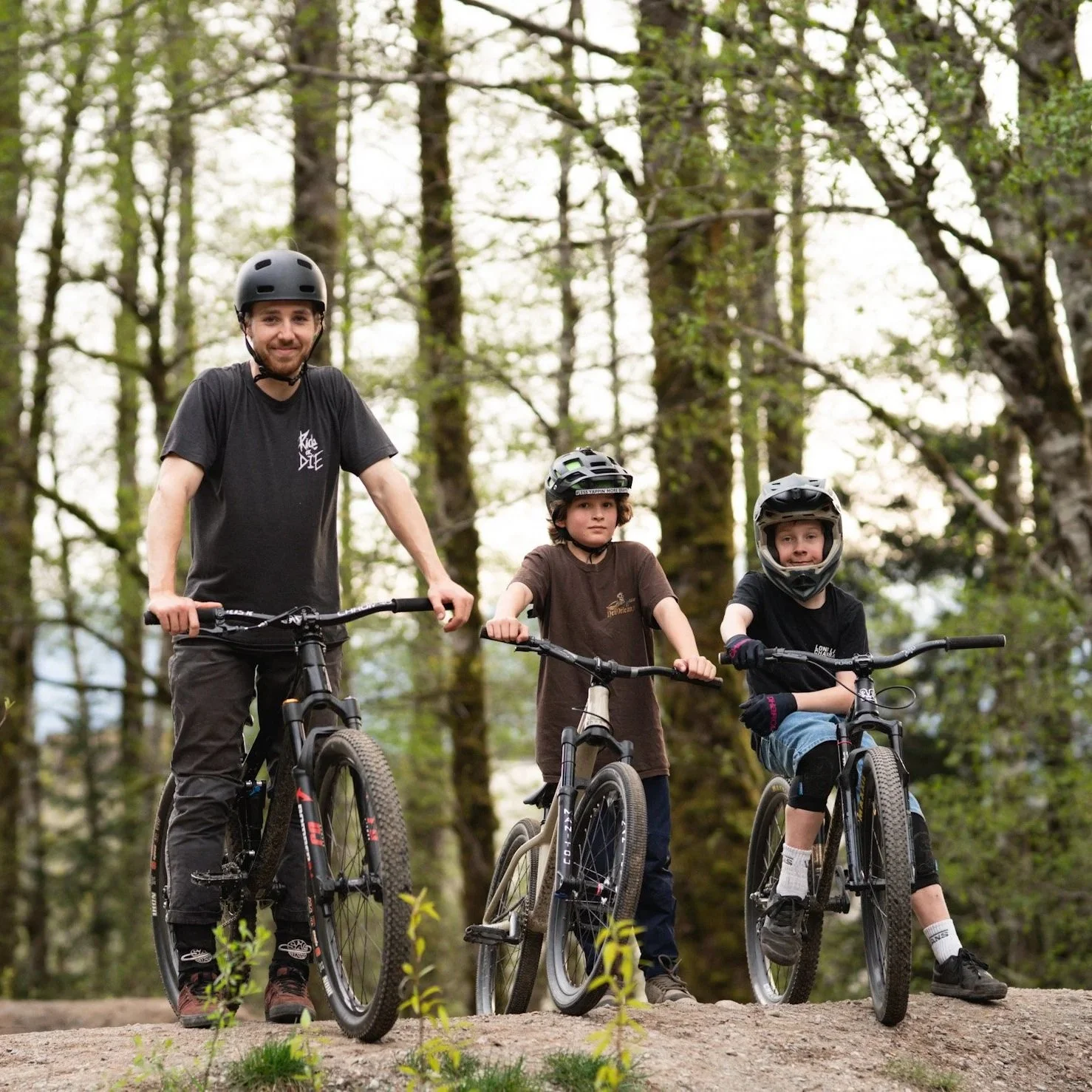 A man and two children on mountain bikes in a forest, all wearing helmets and casual clothing, with greenery and trees in the background.